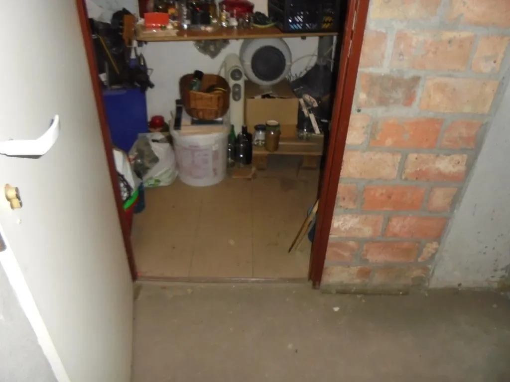 Widok na pomieszczenie magazynowe za otwartymi drzwiami. Po prawej stronie ceglana ściana, po lewej biała drzwi z klamką i zamkiem. Wewnątrz na półkach znajdują się różne przedmioty: pojemniki, wentylator, kosz, wiadro, butelki oraz karton. Podłoga w pomieszczeniu wyłożona jest płytkami, przed drzwiami betonowa podłoga.