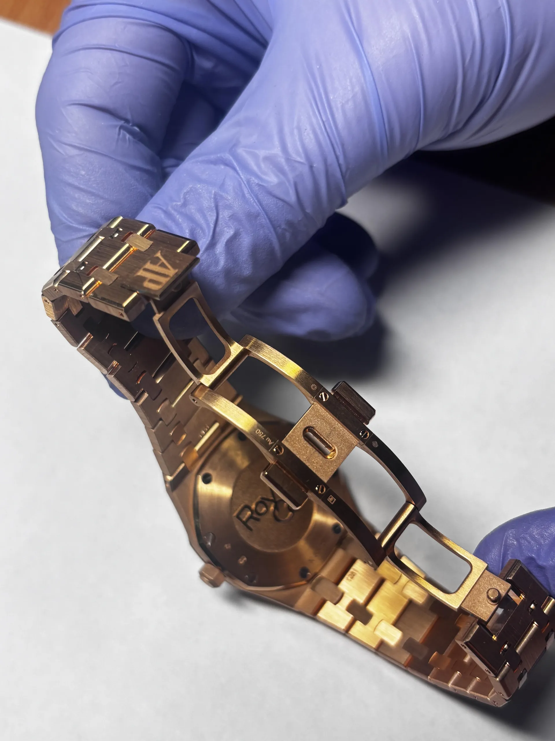 Ręka w niebieskiej rękawiczce trzyma różowozłoty zegarek Rolex, widoczna tylna część z wygrawerowanym napisem 'ROLEX' oraz klamra z napisem 'AP' na bransolecie. Tło stanowi jasna powierzchnia.