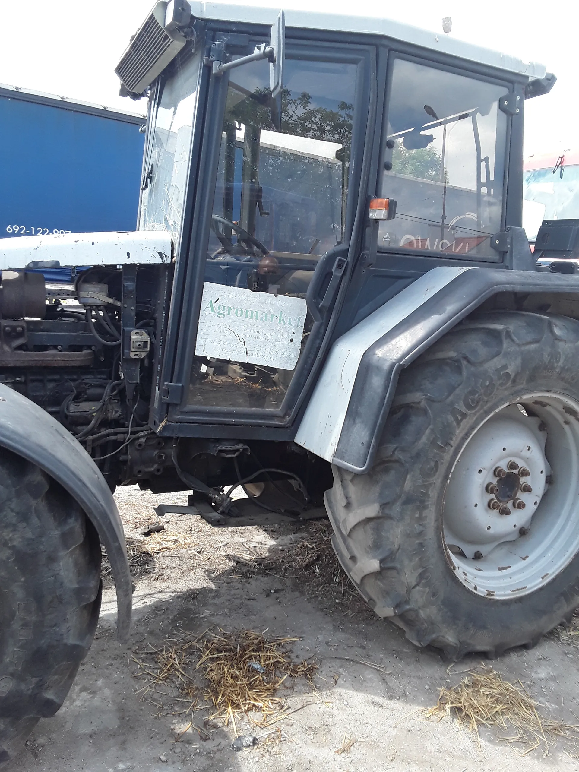 Zbliżenie na ciągnik rolniczy. Widoczna kabina z szybami, duże koło z białym felgiem i oponą z widocznym napisem "AGRICULTURAL". Na przedniej szybie kabiny umieszczona tablica z napisem "Agromarket". W tle niebieska tablica z białym tekstem "692-122-007". Podłoże ziemne z suchą trawą.