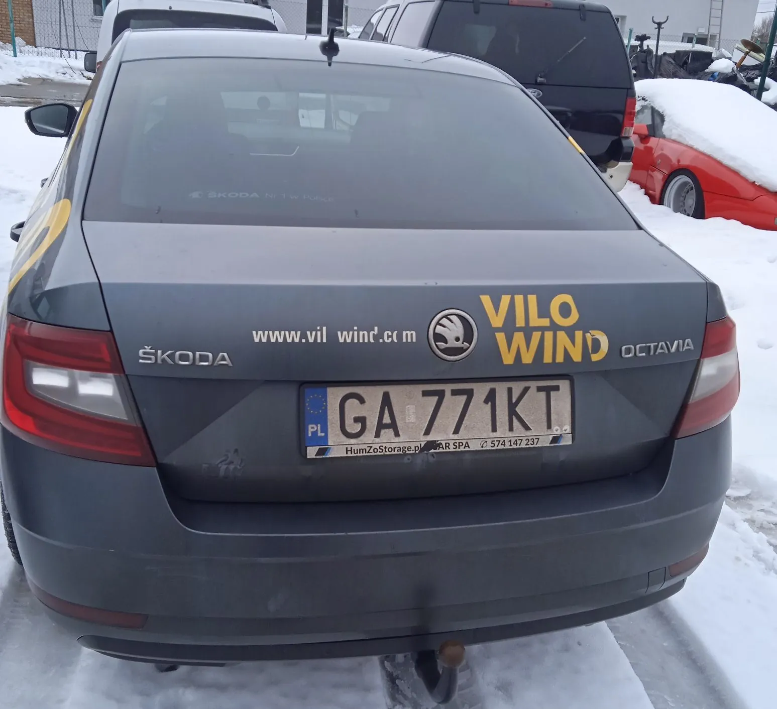 Tylny widok ciemnoszarego samochodu marki Škoda Octavia zaparkowanego na śniegu. Na klapie bagażnika znajdują się napisy: 'ŠKODA' po lewej stronie, 'OCTAVIA' po prawej oraz żółty napis 'VILO WIND' wraz z adresem strony internetowej 'www.vilwind.com' i logo marki. Samochód posiada polską tablicę rejestracyjną o numerze 'GA 771KT'. Poniżej tablicy widnieje ramka z tekstem 'HumZoStorage.pl' oraz numerem telefonu '574 147 237'. W tle widoczne są inne pojazdy przykryte śniegiem.