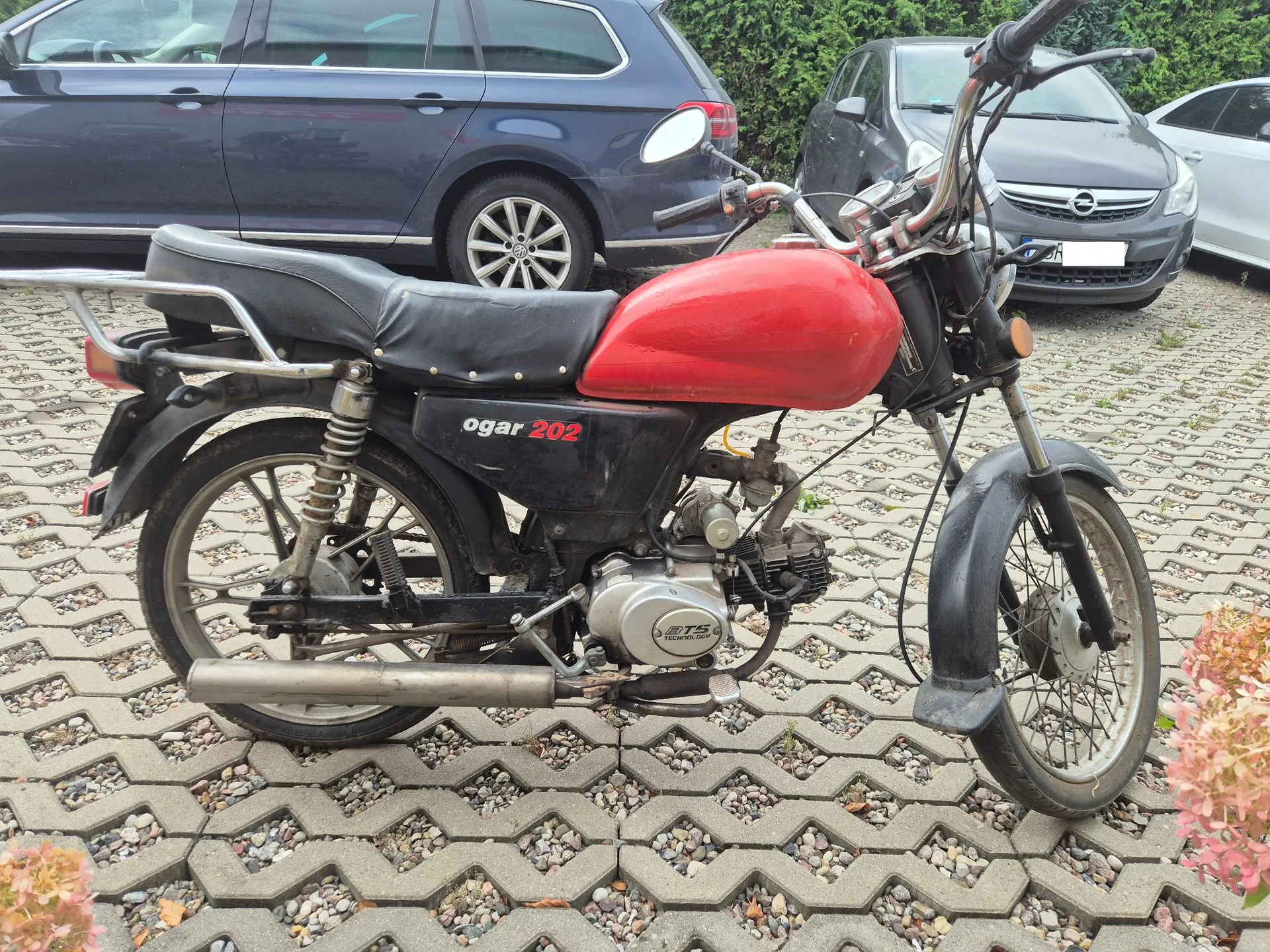 Czerwono-czarny motocykl marki Ogar 202 zaparkowany na kostce brukowej. Pojazd ma czerwony zbiornik paliwa, czarną kanapę i czarne błotniki. Na boku ramy widnieje napis 'ogar 202'. W tle widoczne są zaparkowane samochody: granatowy kombi po lewej oraz srebrny samochód osobowy po prawej.