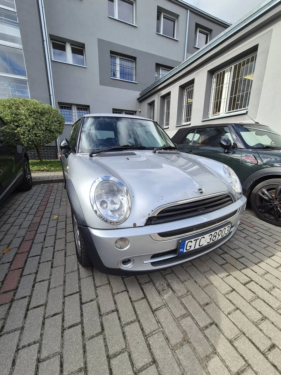 Srebrny samochód Mini Cooper zaparkowany na kostce brukowej przed szarym, wielopiętrowym budynkiem. Samochód ma widoczne uszkodzenia lakieru na masce i zderzaku. Tablica rejestracyjna samochodu to GTC 38903. Po prawej stronie widoczny jest fragment ciemnozielonego samochodu Mini.