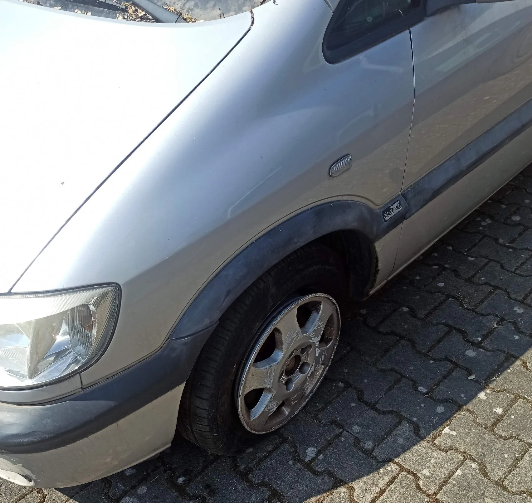 Zbliżenie na przedni lewy bok srebrnego samochodu. Widoczny jest fragment maski, reflektor, przedni błotnik oraz koło z aluminiową felgą. Samochód stoi na kostce brukowej.