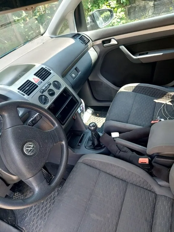 Wnętrze samochodu marki Volkswagen, widok z perspektywy kierowcy. Widoczna jest czarna kierownica z logo marki, deska rozdzielcza z panelami sterowania klimatyzacją i radiem, lewy fotel pasażera oraz drzwi od strony pasażera. Tapicerka foteli jest ciemnoszara w delikatną kratkę.