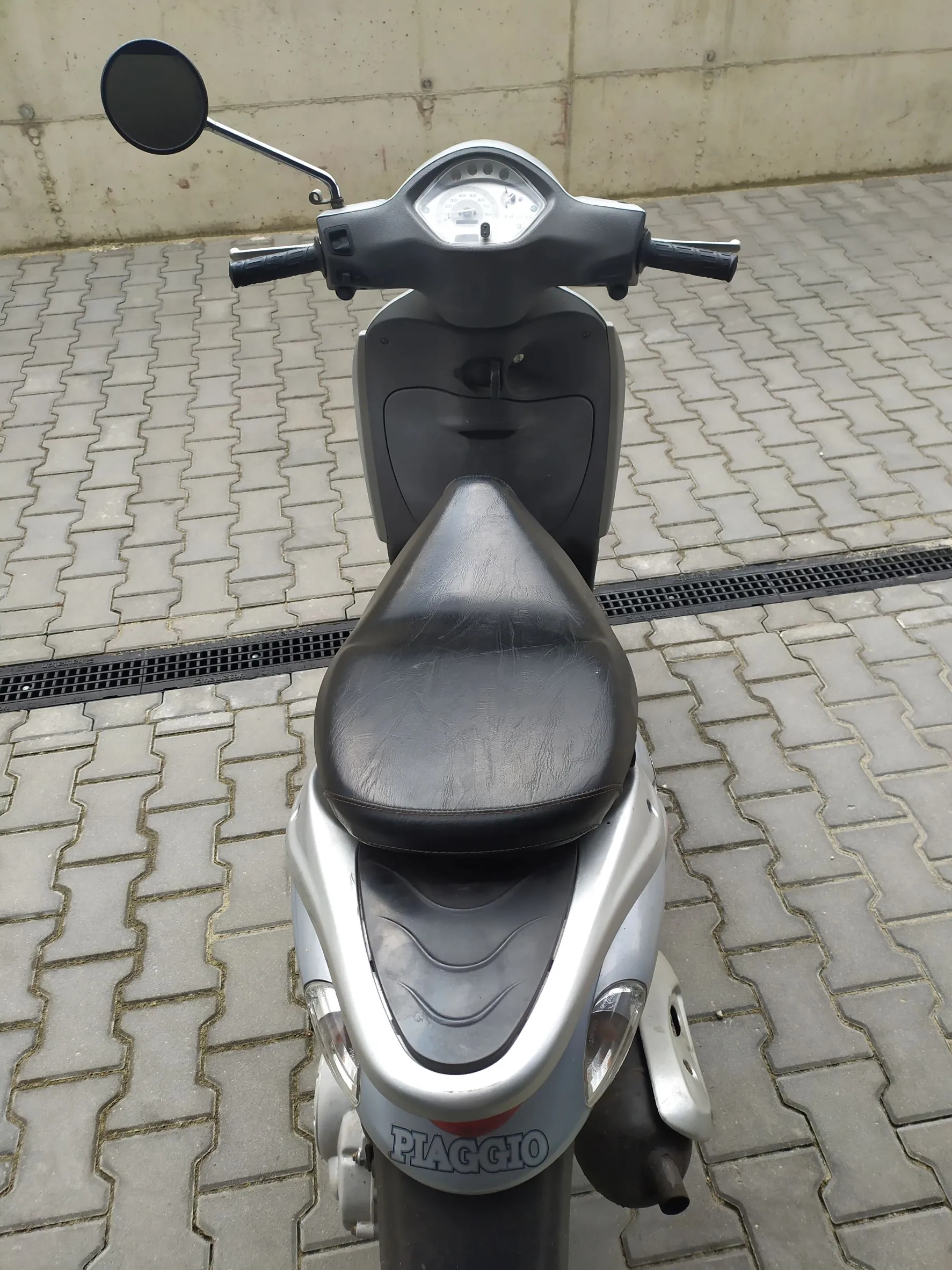 Srebrno-szary skuter marki Piaggio zaparkowany na kostce brukowej. Widoczny jest profil pojazdu z czarnym siedzeniem, kierownicą z lusterkiem oraz napisem 'PIAGGIO' na tylnej obudowie.