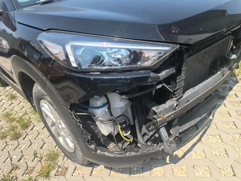 Zbliżenie na uszkodzony przód czarnego samochodu. Brakuje przedniego zderzaka i części maski, co odsłania wewnętrzne elementy konstrukcyjne, w tym chłodnicę oraz biały plastikowy zbiornik wyrównawczy z podłączonymi niebieskim i żółtym przewodem. Widoczny jest prawy reflektor oraz przednie koło z felgą aluminiową. Samochód stoi na kostce brukowej z otworami na trawę.