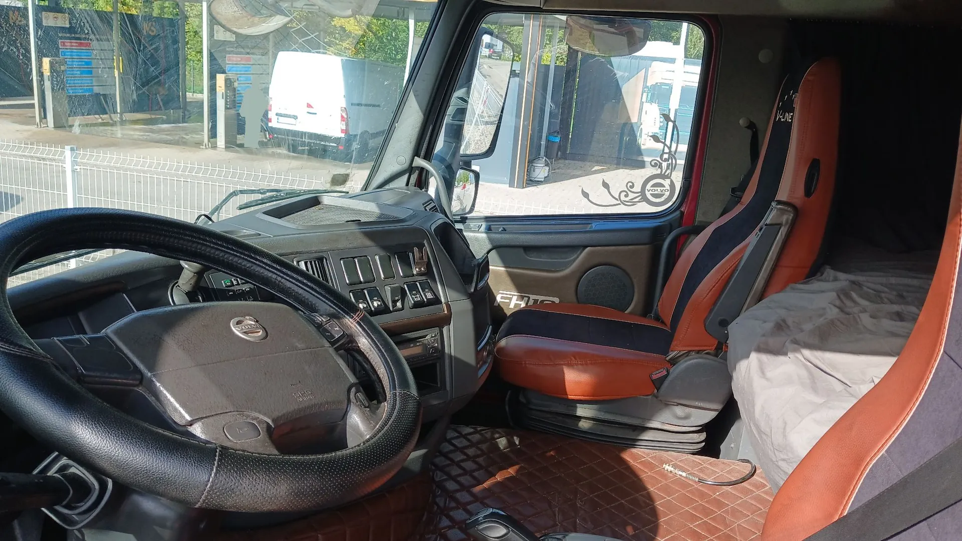 Wnętrze kabiny ciężarówki marki Volvo. Widoczna jest czarna kierownica z logo Volvo, deska rozdzielcza z licznymi przełącznikami i przyciskami oraz brązowy fotel kierowcy z czarnymi wstawkami. W tle, przez przednią szybę, widoczny jest biały samochód dostawczy zaparkowany na zewnątrz. Podłoga kabiny jest wyłożona brązową matą o strukturze rombów.