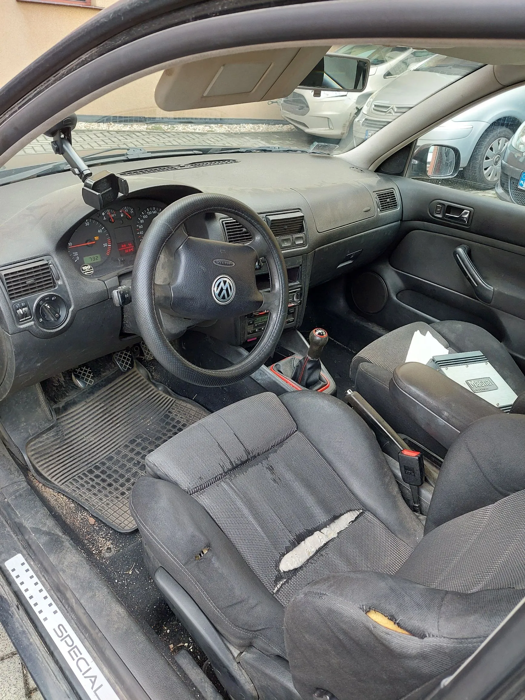 Wnętrze samochodu widziane z perspektywy otwartych drzwi kierowcy. Widoczna jest czarna deska rozdzielcza, kierownica z logo Volkswagena, zegary oraz dwa przednie fotele z czarnej tkaniny. Fotel kierowcy jest mocno zużyty i posiada widoczne rozdarcia na siedzisku i oparciu. Na podłodze znajduje się gumowa mata. Na progu samochodu widnieje napis 'SPECIAL'. Przez przednią szybę widać zaparkowane inne samochody.