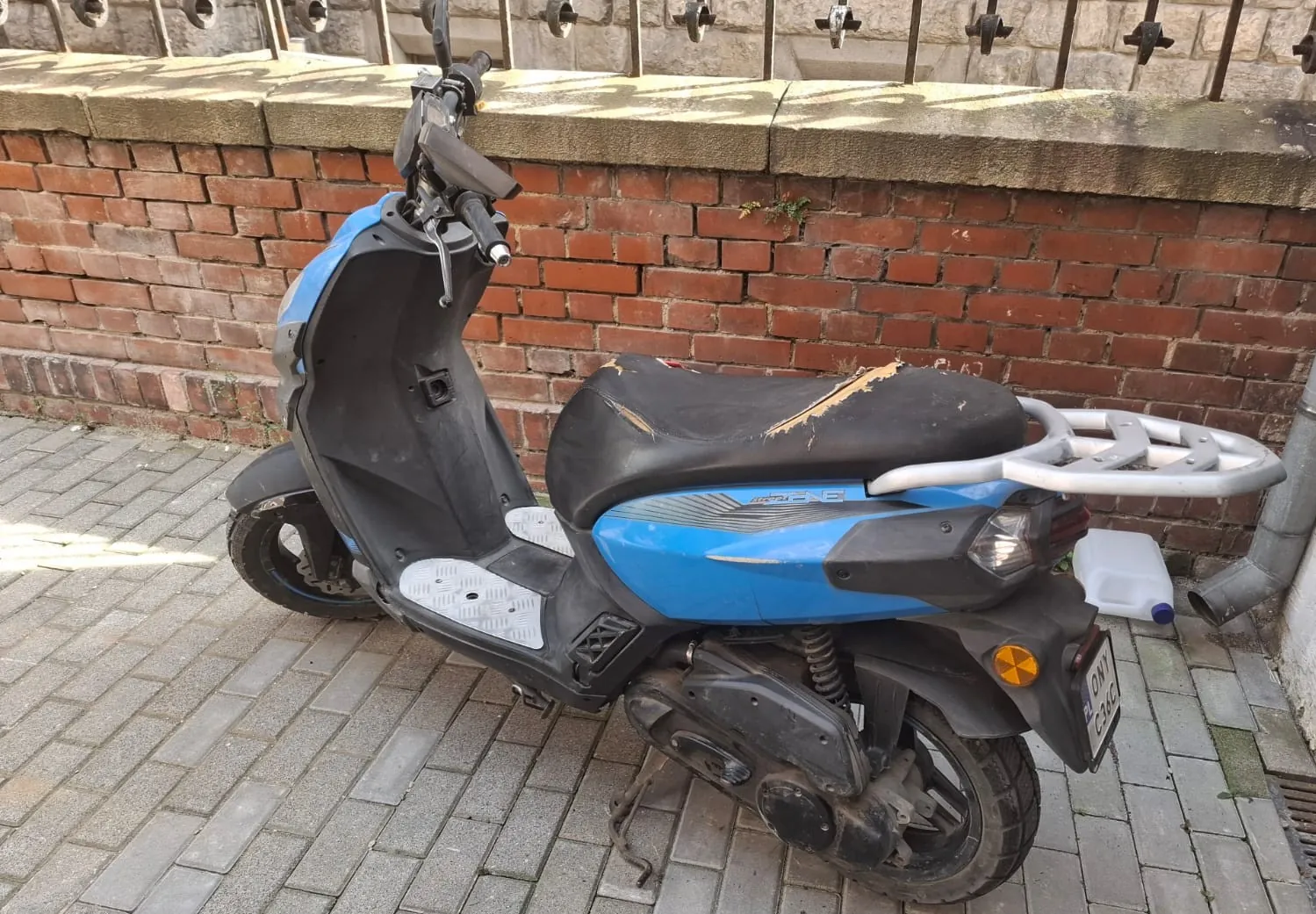 Niebiesko-czarny skuter zaparkowany na kostce brukowej przy czerwonej ceglanej ścianie. Pojazd ma widoczne uszkodzenia w postaci rozdarcia na czarnym siedzeniu. Z tyłu znajduje się srebrny bagażnik oraz tablica rejestracyjna. W tle widać betonowy murek zwieńczony metalowym ogrodzeniem.