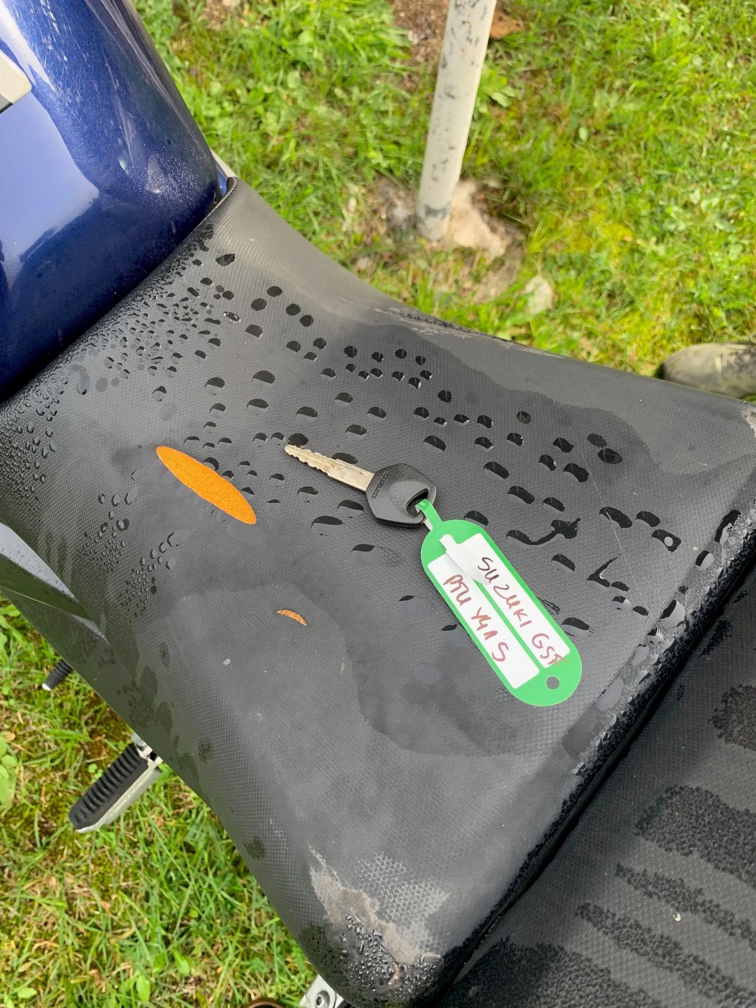 Zbliżenie na mokre, czarne siedzenie motocykla, na którym leży kluczyk z zielonym brelokiem. Na breloku znajduje się odręczny napis: 'SUZUKI GST DTU Y415'. Na siedzeniu widoczne są krople wody oraz mała, pomarańczowa plama. W tle widać zieloną trawę i fragment białego słupka.