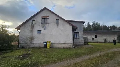 Zdjęcie przedstawia stary, dwukondygnacyjny budynek o szarym tynku z dwuspadowym dachem. Na ścianie widoczne są niewielkie okna oraz ślady wilgoci. Przed budynkiem stoją dwa pojemniki na odpady: jeden czarny i jeden żółty. Po prawej stronie znajduje się niższa, przylegająca zabudowa gospodarcza. W oddali, po prawej stronie, stoi postać człowieka. Całość znajduje się na trawiastym terenie z widoczną drogą gruntową, w otoczeniu drzew pod zachmurzonym niebem.