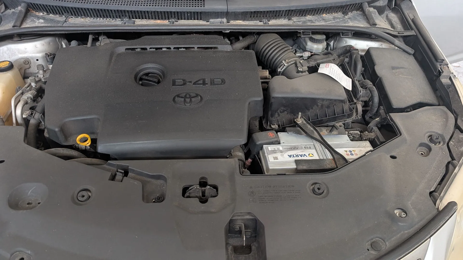 Widok z góry na komorę silnika samochodu marki Toyota. Centralnie znajduje się czarna plastikowa osłona silnika z napisem „D-4D” oraz logo Toyoty. Po prawej stronie widoczny jest akumulator marki VARTA oraz inne elementy układu elektrycznego i mechanicznego. Całość jest zakurzona.