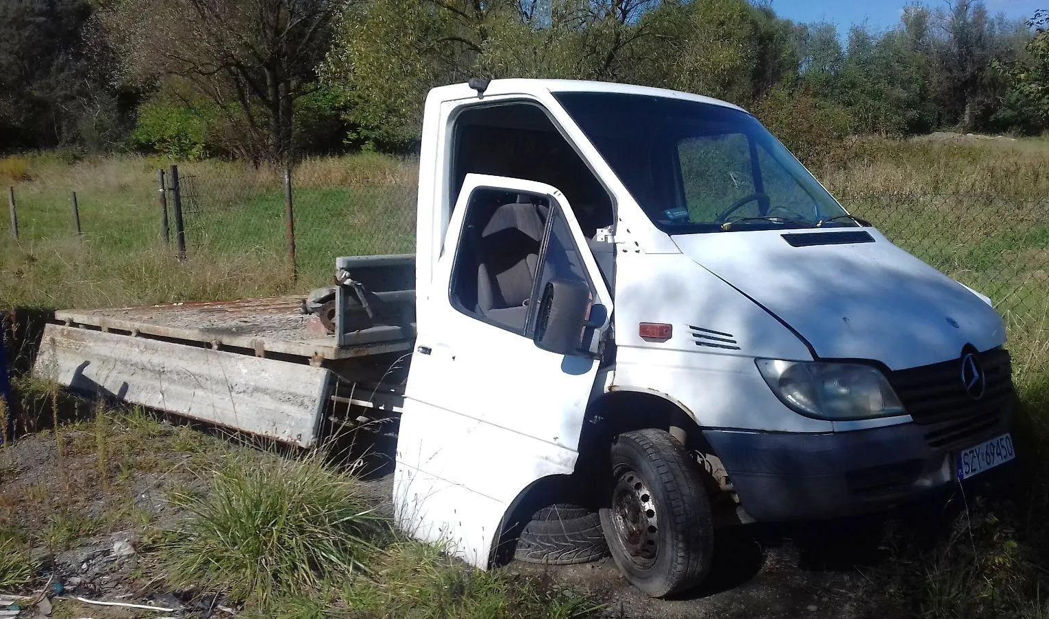 Biały samochód dostawczy marki Mercedes-Benz z otwartymi drzwiami kierowcy, zaparkowany na trawiastym terenie. Pojazd posiada otwartą platformę ładunkową z tyłu. Na przednim zderzaku widoczna jest tablica rejestracyjna o numerze SZY 694S0. W tle znajdują się drzewa i krzewy pod jasnym niebem.