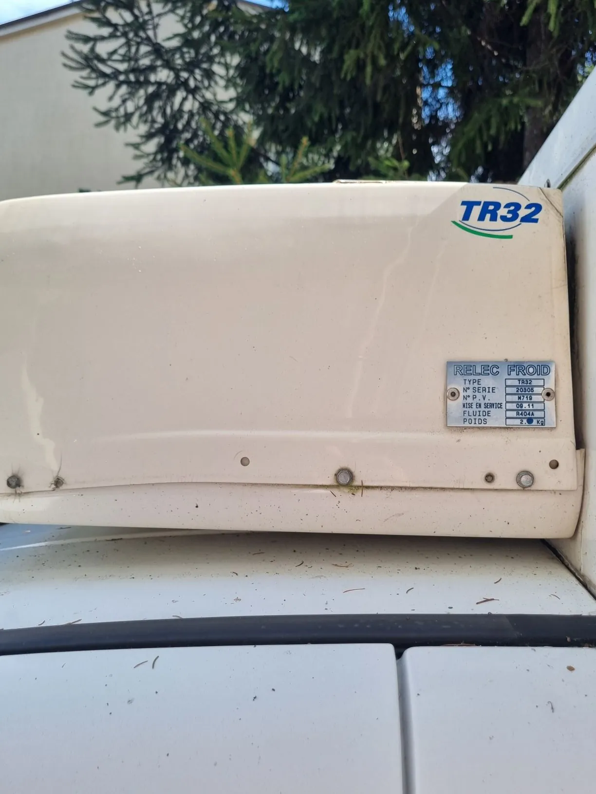 Zbliżenie na białą obudowę urządzenia chłodniczego zamontowanego na dachu pojazdu. W prawym górnym rogu znajduje się niebieski napis 'TR32'. Po prawej stronie widoczna jest tabliczka znamionowa z napisem 'RELEC FROID' oraz danymi technicznymi: TYPE TR32, N° SERIE 20306, N° P.T. M716, DATE DE SERVICE 06-11-11, FLUIDE R404A, POIDS 2,1 kg.