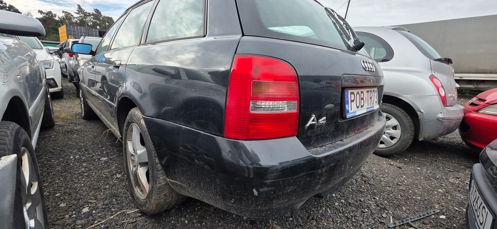 Czarny samochód marki Audi A4 w nadwoziu kombi, zaparkowany na żwirowym placu wśród innych pojazdów. Widoczny jest tył auta z prawym tylnym światłem, logo Audi, oznaczeniem modelu A4 oraz tablicą rejestracyjną o numerze POB TR73. Samochód jest zakurzony i posiada widoczne zarysowania na tylnym zderzaku.