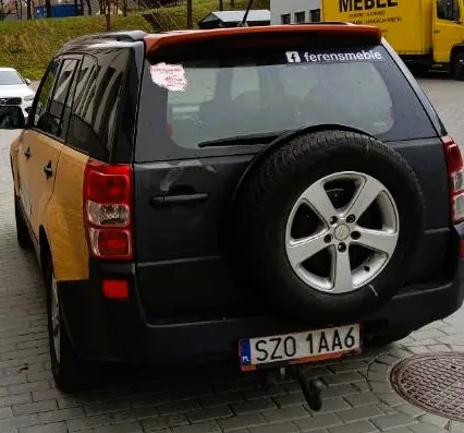 Tylny widok ciemnoszarego samochodu typu kombi z zapasowym kołem zamocowanym na klapie bagażnika. Samochód ma żółte elementy nadwozia na bokach. Na tylnej szybie znajduje się napis 'f ferensmeble' oraz czerwona naklejka. Tablica rejestracyjna ma numer SZO 1AA6.