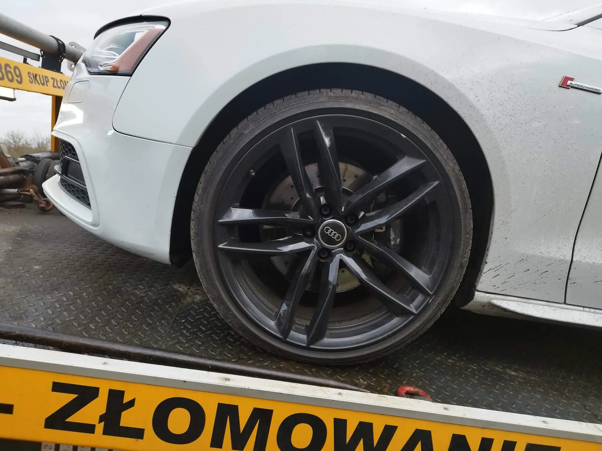 Zbliżenie na przednie lewe koło srebrnego samochodu marki Audi. Samochód posiada czarną felgę aluminiową z pięcioma podwójnymi ramionami i logo Audi w centrum. Pojazd znajduje się na platformie z metalową, ryflowaną powierzchnią. W dolnej części obrazu widoczny jest fragment żółtego napisu „ZŁOMOWANIE”. W tle po lewej stronie widać fragment żółtej konstrukcji z napisem „69 SKUP ZŁO”.