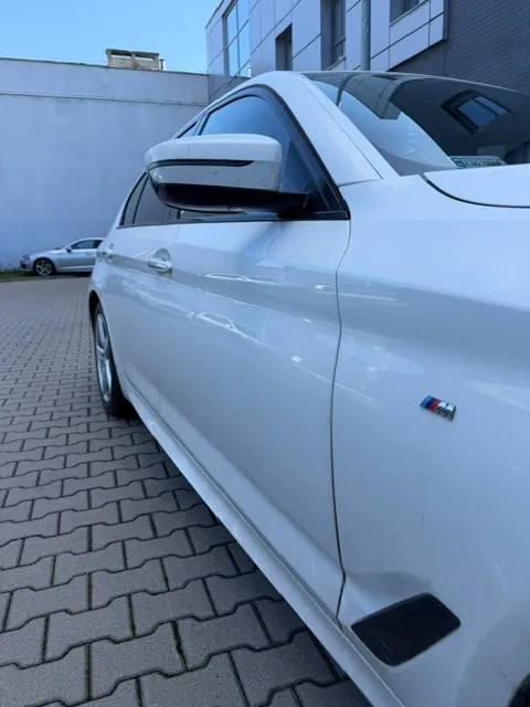 Zbliżenie na prawy bok białego samochodu marki BMW, zaparkowanego na kostce brukowej. Widoczny jest fragment drzwi z logo M, lusterko boczne oraz fragment przedniego koła. W tle widać fragment innego, srebrnego samochodu oraz jasną ścianę budynku.