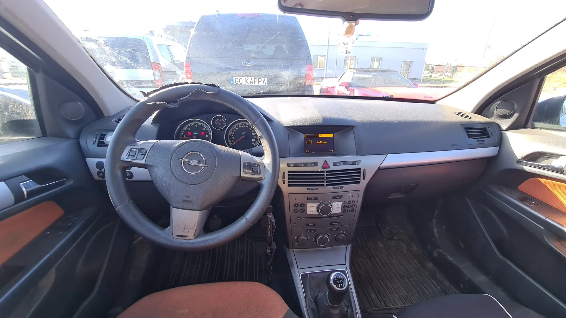 Wnętrze samochodu z widokiem na kierownicę z logo Opel, zniszczoną obudową kierownicy, zegary przyrządów, centralną konsolę z radiem wyświetlającym napis No Phone, dźwignią zmiany biegów oraz widok przez przednią szybę na ciemny samochód Ford z tablicą rejestracyjną GO KAPPA, czerwony samochód i budynek w tle. Wnętrze posiada brązowe i czarne elementy wykończenia.