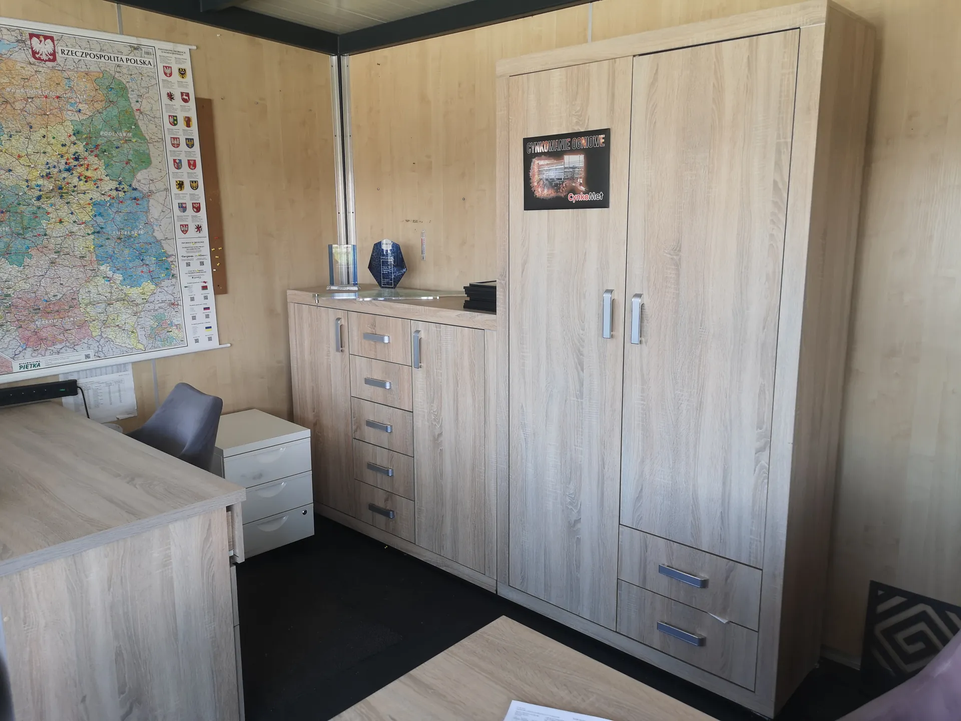 Wnętrze biura z meblami w kolorze jasnego drewna. Po lewej stronie duża mapa Polski z napisem "RZECZPOSPOLITA POLSKA" i godłem orła. Obok mapy szafka z szufladami i blat, na którym znajdują się przezroczysty obiekt, niebieski geometryczny przedmiot oraz stos czarnych dokumentów. Po prawej wysoka szafa z dwiema drzwiami i szufladą, na której umieszczono plakat z napisem "CYNKOWANIE OGNIOWE" oraz "CynkMet" pod zdjęciem. W tle drewniane ściany, czarne podłoga i fragment biurka z krzesłem. Widoczne również białe szuflady obok biurka oraz czarno-biały wzór na podłodze po prawej stronie.
