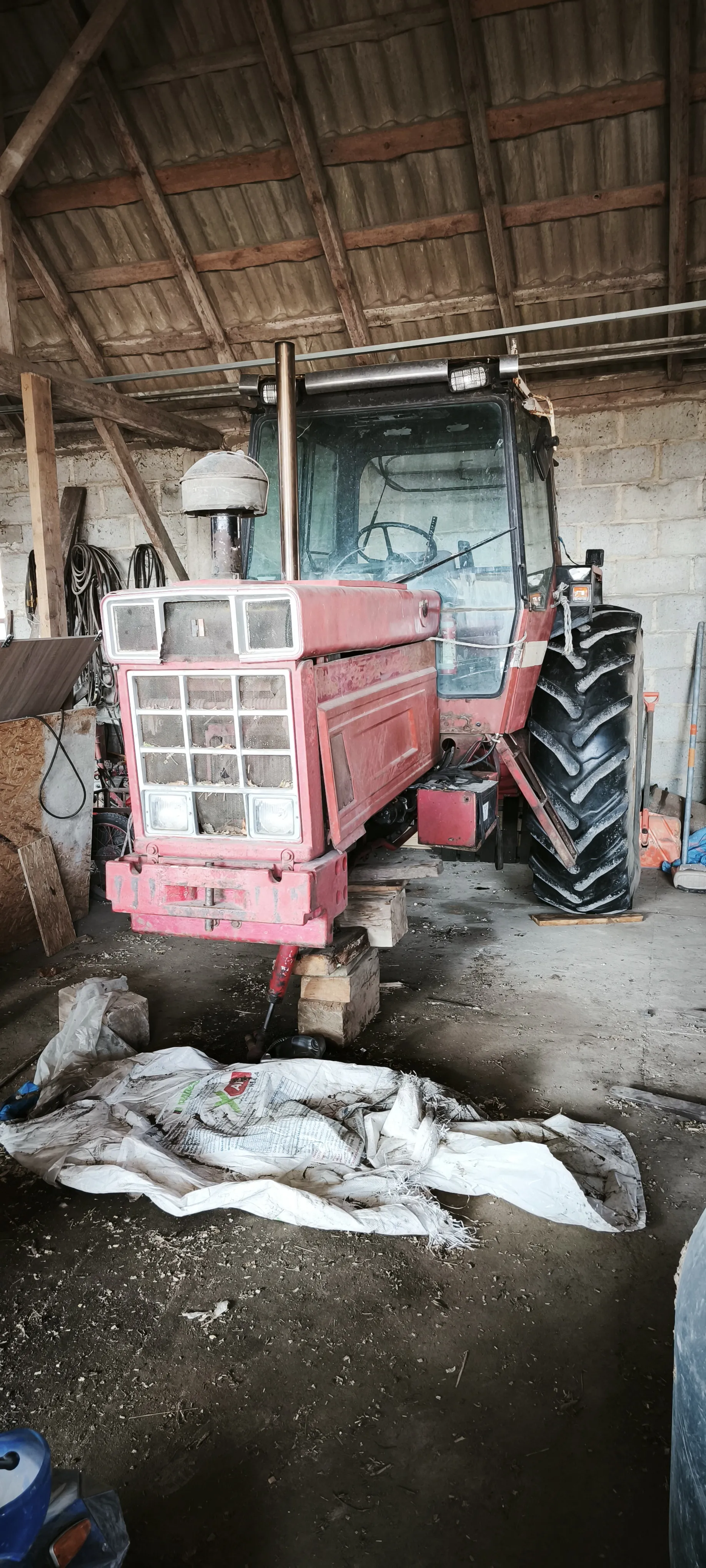 Czerwony traktor w stodole, ustawiony na drewnianych blokach pod przodem. W tle widoczne betonowe bloki ściany, drewniane belki dachowe i narzędzia. Na podłodze leży biały workowaty materiał z nieczytelnym nadrukiem. Duże opony traktora, przednia część z siatką i reflektorami. W lewym dolnym rogu fragment niebieskiego obiektu.