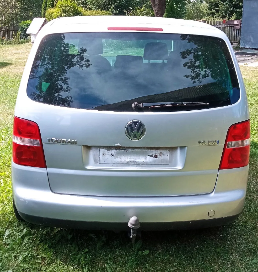 Tylny widok srebrnego samochodu marki Volkswagen Touran. Na klapie bagażnika widoczne jest logo Volkswagena, napis 'TOURAN' po lewej stronie oraz oznaczenie silnika '1.6 FSI' po prawej stronie. Samochód posiada hak holowniczy i jest zaparkowany na trawie. W tle widoczna jest zieleń i fragment ogrodzenia.