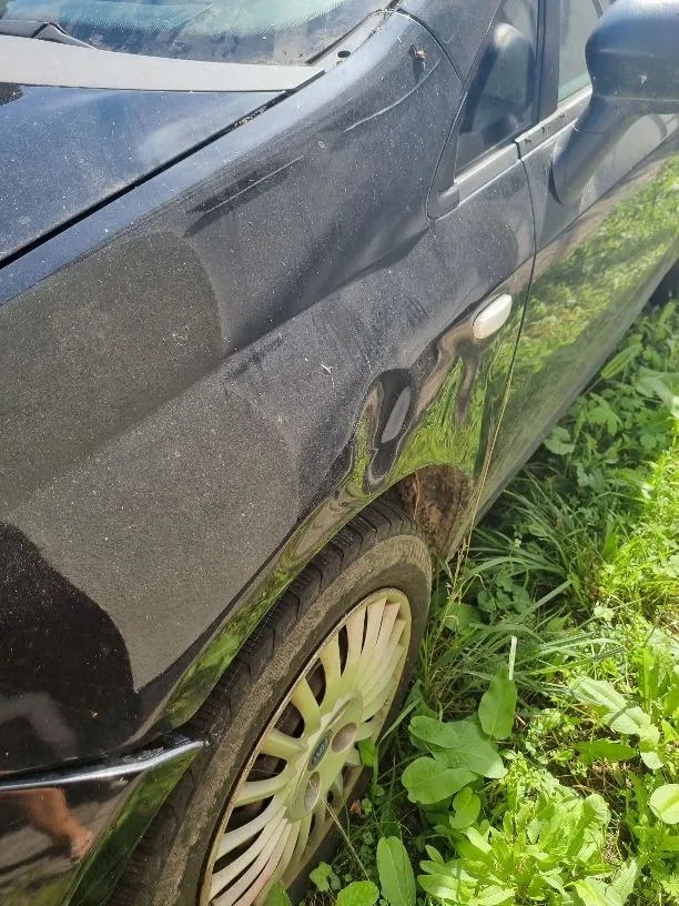 Zbliżenie na przedni lewy błotnik i koło czarnego samochodu. Karoseria jest zakurzona, a nad nadkolem widoczne są ślady korozji i ubytki lakieru. Samochód stoi na trawie.