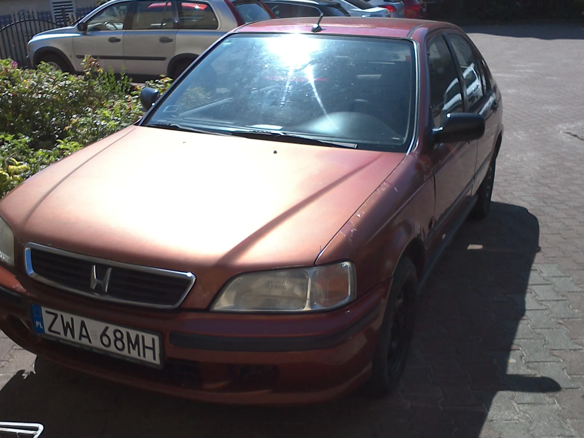 Pomarańczowy samochód osobowy marki Honda zaparkowany na kostce brukowej. Widoczna jest przednia część pojazdu z tablicą rejestracyjną o treści ZWA 68MH. W tle znajduje się inny, srebrny samochód oraz zielone krzewy.