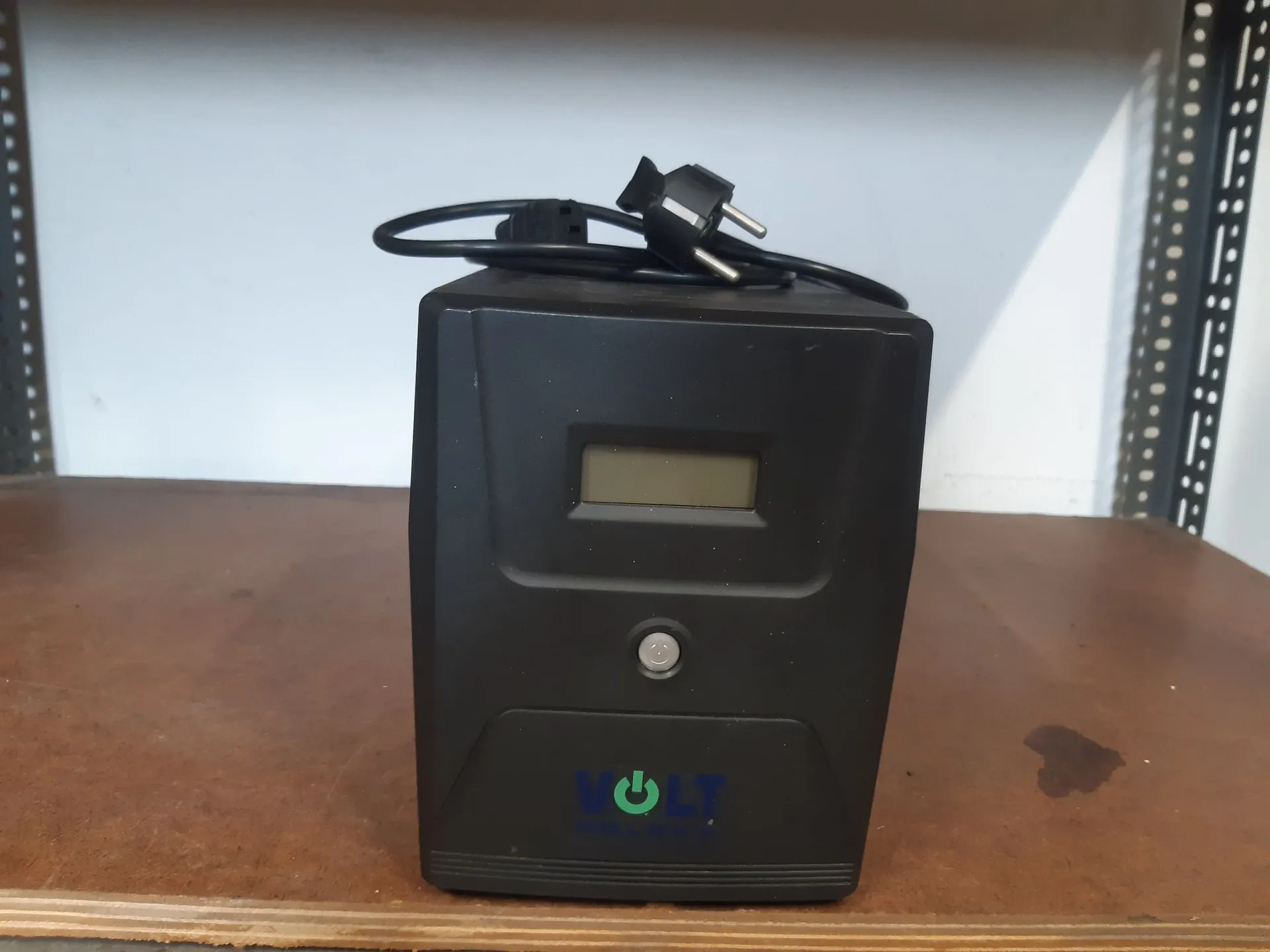 Czarny, prostokątny zasilacz lub urządzenie elektroniczne stojące na brązowej powierzchni. Urządzenie posiada mały, prostokątny wyświetlacz LCD w górnej części, okrągły przycisk poniżej oraz zielone logo z napisem 'VOLT' na dole. Z góry urządzenia wychodzi czarny kabel z wtyczką sieciową typu C.