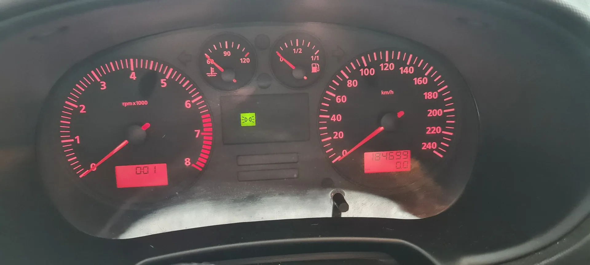Panel instrumentów samochodu z czerwonym podświetleniem. Po lewej znajduje się obrotomierz z oznaczeniem 'rpm x 1000', w centrum dwa mniejsze wskaźniki (temperatury cieczy chłodzącej i poziomu paliwa) oraz zielona kontrolka, a po prawej prędkościomierz z oznaczeniem 'km/h'. Na dole prędkościomierza znajduje się cyfrowy licznik kilometrów wyświetlający wartość 184699.9.