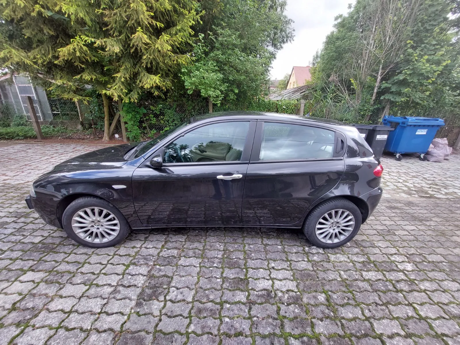 Czarny samochód osobowy typu hatchback zaparkowany bokiem na kostce brukowej z mchem w szczelinach. W tle widoczne są gęste zielone drzewa i krzewy oraz niebieski kontener na odpady po prawej stronie.