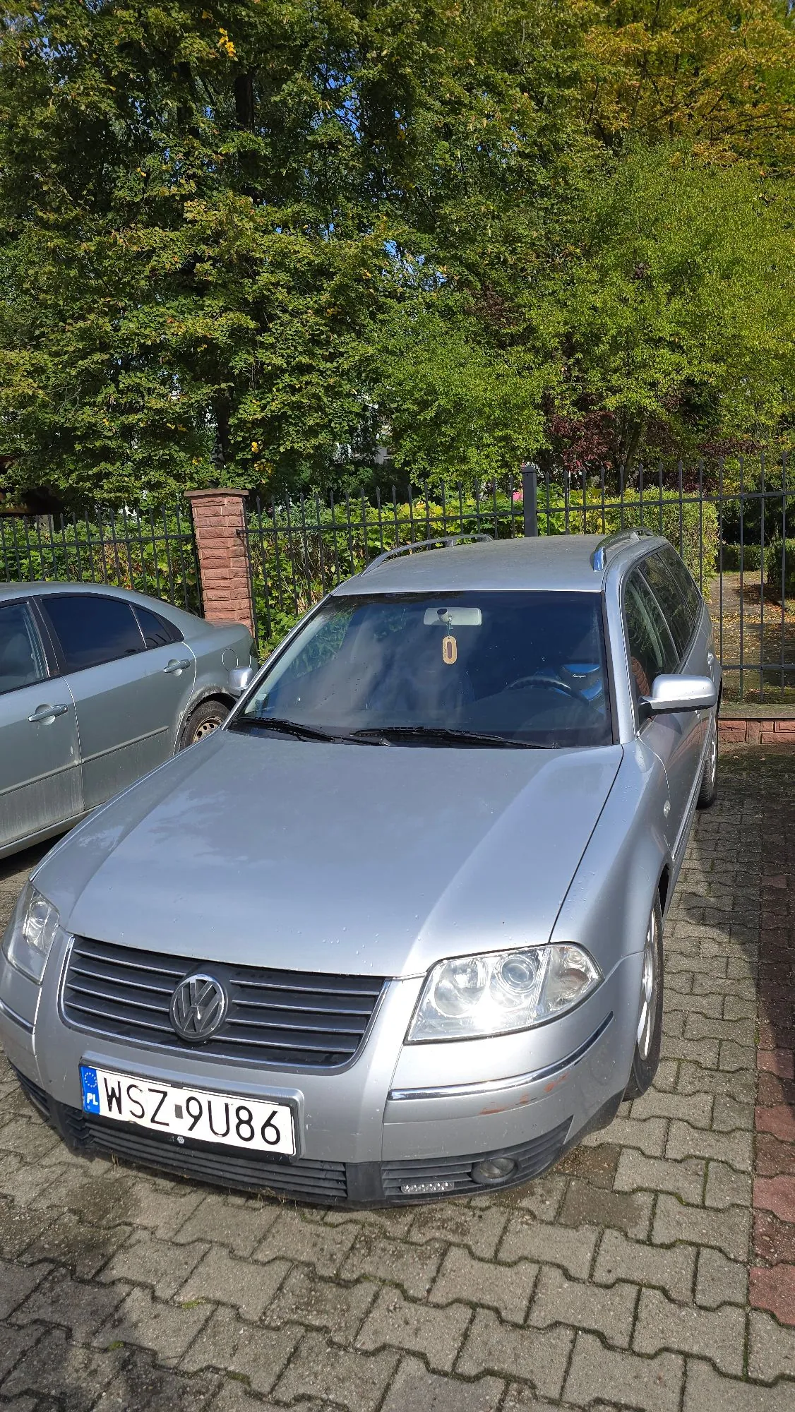 Srebrny samochód marki Volkswagen w nadwoziu kombi zaparkowany na kostce brukowej. Na przedniej szybie widoczna jest żółta naklejka z cyfrą 0. Samochód posiada polską tablicę rejestracyjną o numerze WSZ 9U86. W tle znajduje się czarny metalowy płot, ceglany słupek oraz gęste, zielone drzewa.