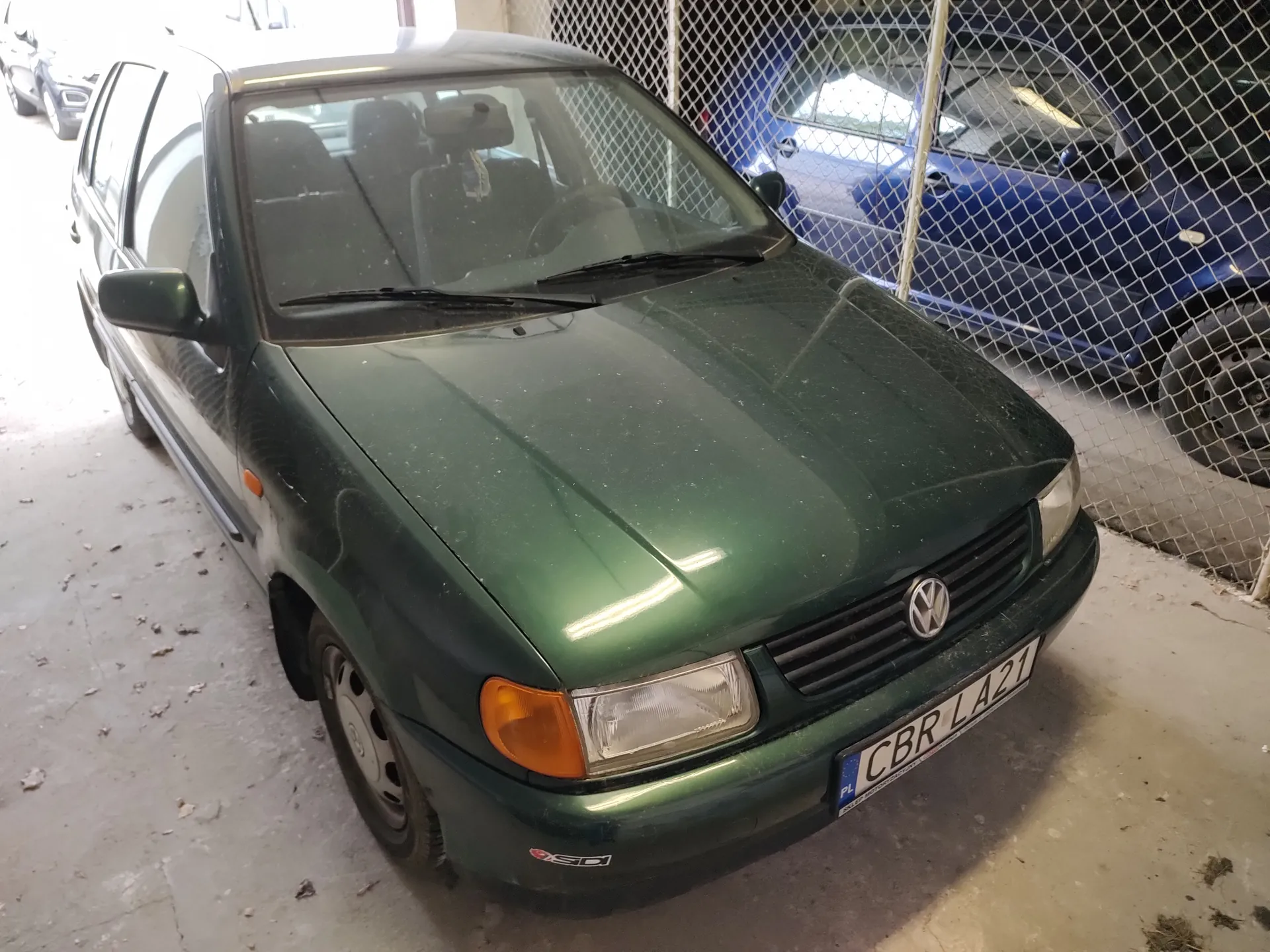 Zielony samochód marki Volkswagen zaparkowany na betonowej powierzchni. Widoczna tablica rejestracyjna CBR LA21 z oznaczeniem PL. W tle niebieski samochód za siatką ogrodzeniową. Na przednim zderzaku zielonego pojazdu znajduje się niewielki naklejony znak. Podłoga pokryta jest drobnymi liśćmi i śmieciem.