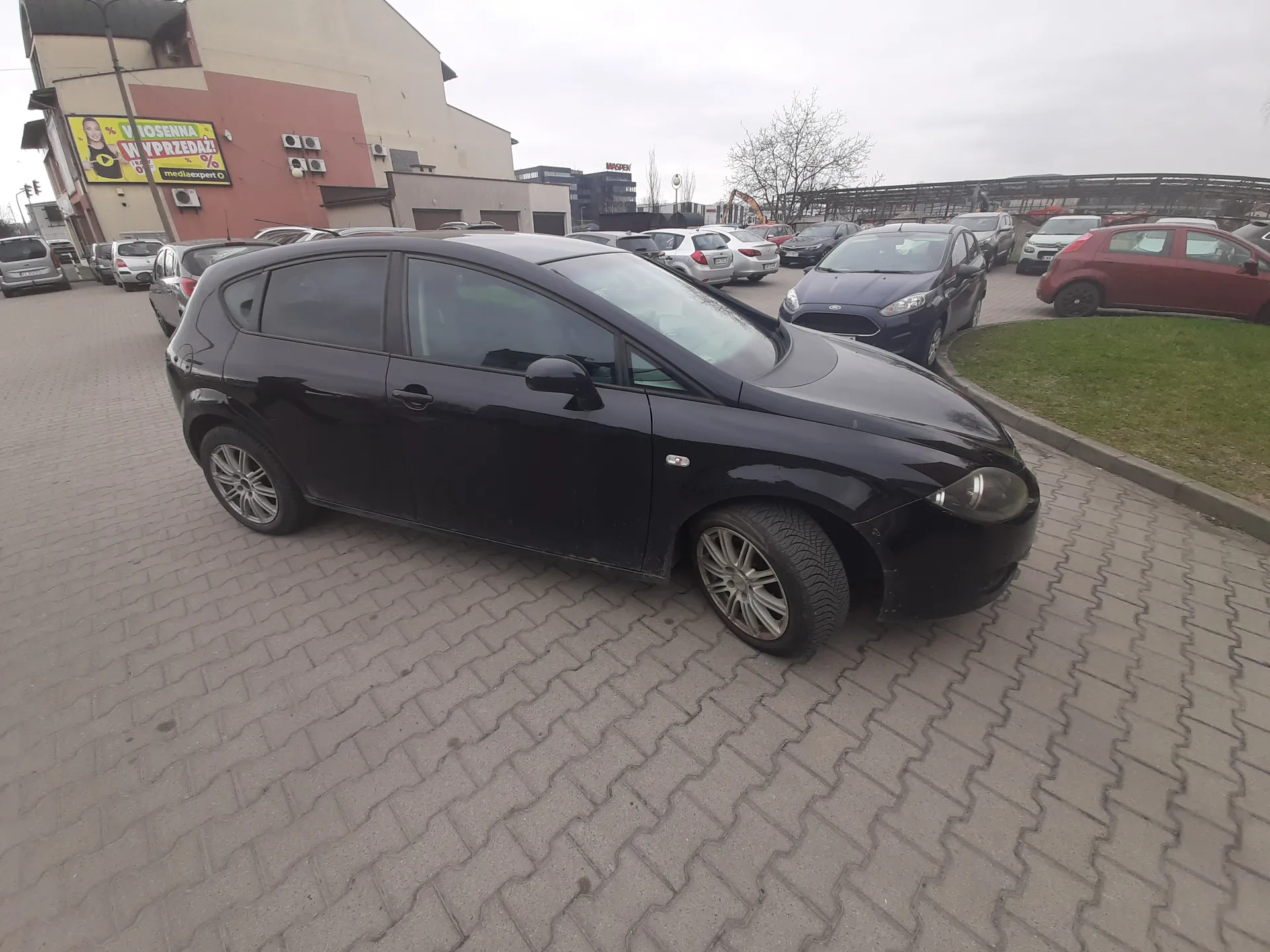 Czarny samochód hatchback zaparkowany na brukowanej powierzchni. W tle inne pojazdy, budynek z żółto-czerwonym banerem reklamowym z napisem 'WYPRZEDAZ!' oraz 'mediaexpert'. Niebo zachmurzone, po prawej stronie trawnik.