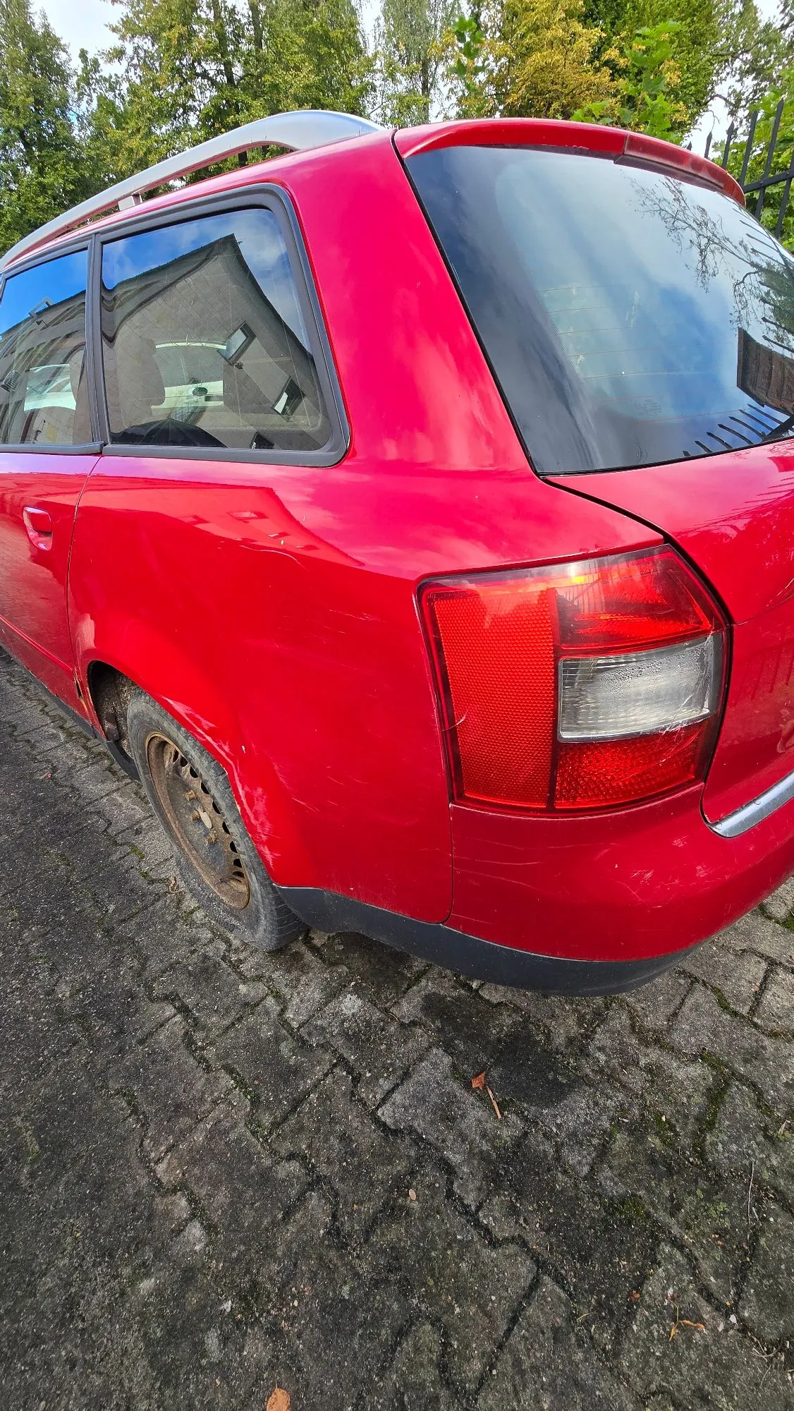 Tylna część czerwonego samochodu typu kombi zaparkowanego na kostce brukowej. Widoczny jest tylny prawy błotnik, lampa tylna oraz fragmenty bocznych szyb i srebrne relingi dachowe. W tle znajdują się zielone drzewa.