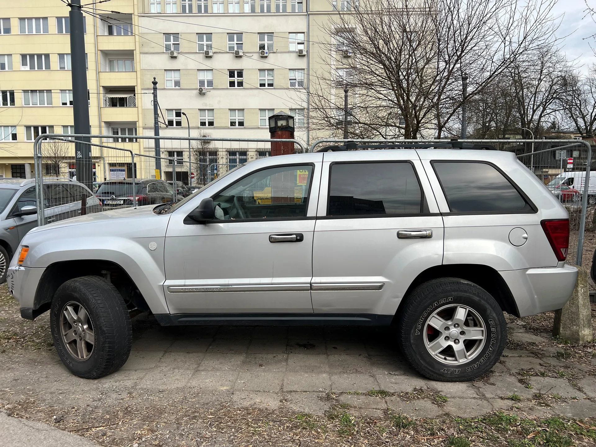 Srebrny SUV typu Jeep Grand Cherokee zaparkowany na betonowej powierzchni. Widok z boku, cztery drzwi, ciemne szyby, dachowy bagażnik. W tle wielopiętrowy budynek z oknami i klimatyzatorami, metalowe ogrodzenie, inne zaparkowane samochody oraz bezlistne drzewa. Opony z widocznymi napisami "Yokohama" i "Geolandar". Niebo zachmurzone.