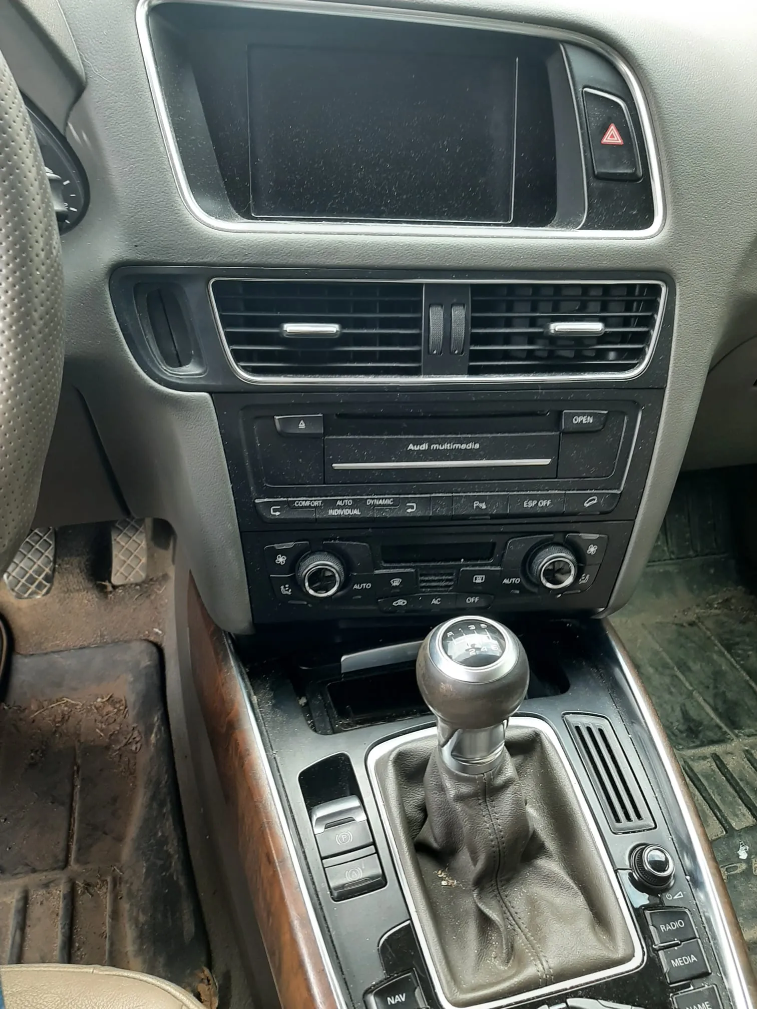 Wnętrze samochodu marki Audi z widocznym panelem środkowym. Na górze znajduje się czarny ekran multimedialny, poniżej wentylacja z metalowymi listwami. Centralnie umieszczony jest odtwarzacz z napisem "Audi multimedia" oraz przyciskiem "OPEN". Poniżej znajdują się przyciski klimatyzacji z oznaczeniami: "COMFORT", "AUTO", "DYNAMIC", "INDIVIDUAL", "ESP OFF" oraz pokrętła z napisami "AUTO" i "AC". W dolnej części widoczna jest dźwignia zmiany biegów z metalową głowicą i skórzaną obudową, a także przyciski oznaczone jako "RADIO", "MEDIA", "NAV". Po lewej stronie fragment kierownicy z perforacją, a na podłodze brudne dywaniki. W prawym górnym rogu przycisk awaryjny z czerwonym trójkątem.