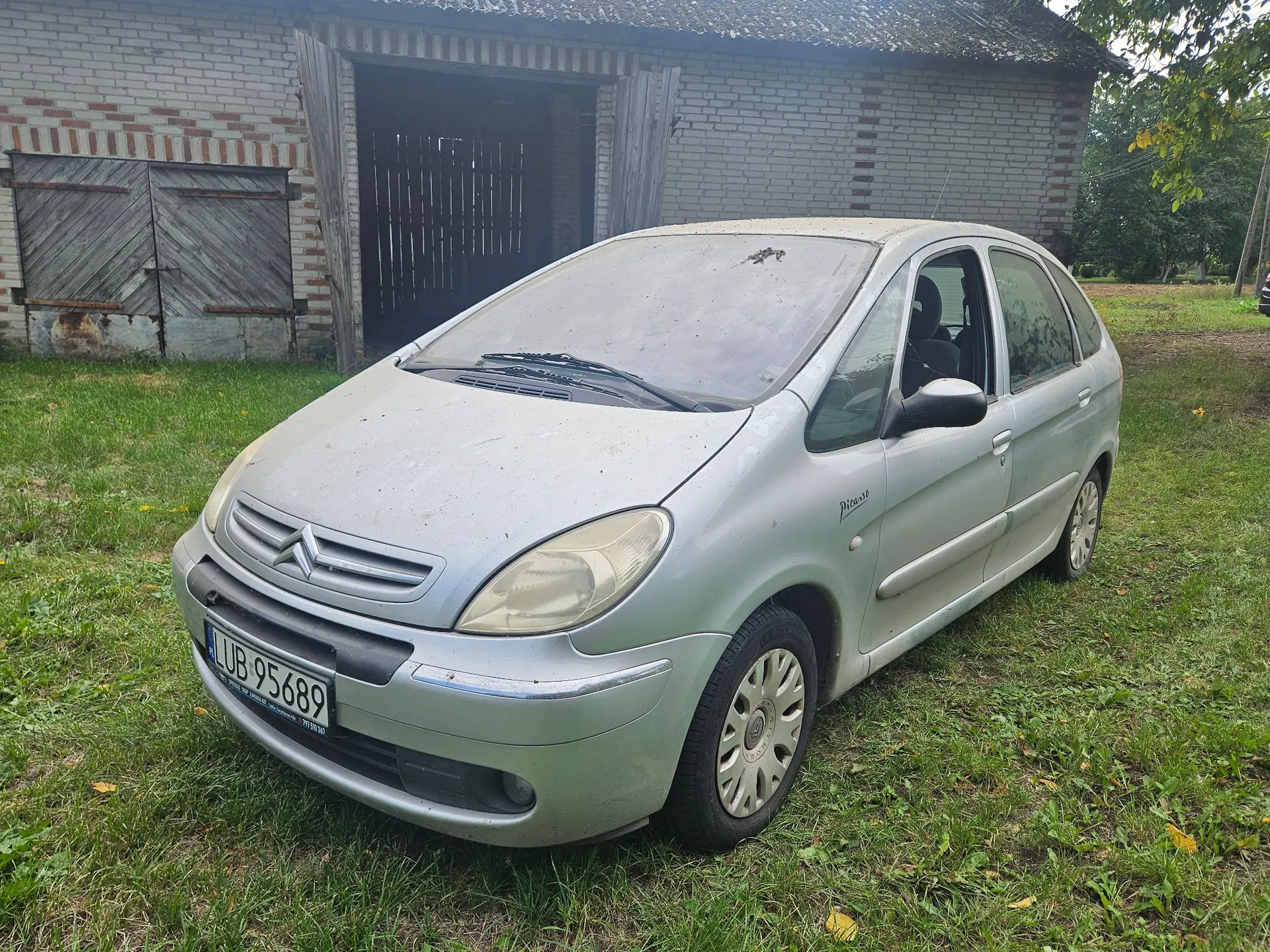 Srebrny samochód marki Citroën Picasso zaparkowany na trawie przed budynkiem z jasnej cegły z drewnianymi drzwiami. Samochód ma tablicę rejestracyjną o numerze LUB 95689.