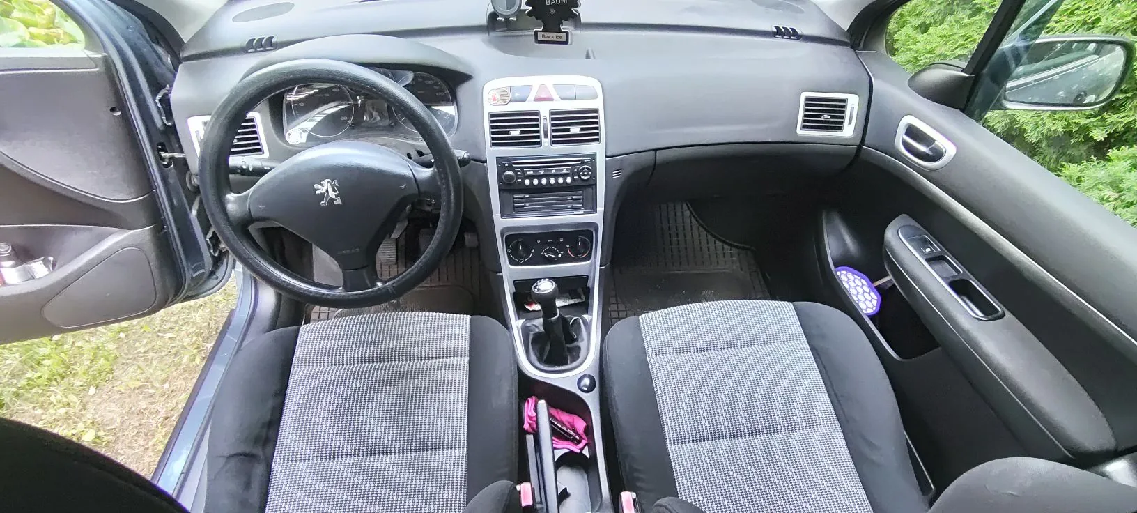 Wnętrze samochodu marki Peugeot, widok z perspektywy pasażera i kierowcy. Widoczna czarna deska rozdzielcza, kierownica z logo lwa, zestaw wskaźników, centralna konsola z radiem i pokrętłami klimatyzacji oraz dwa przednie fotele z szaro-czarną tapicerką w kratkę. Przez przednią szybę i otwarte drzwi widoczna jest zieleń roślinności.