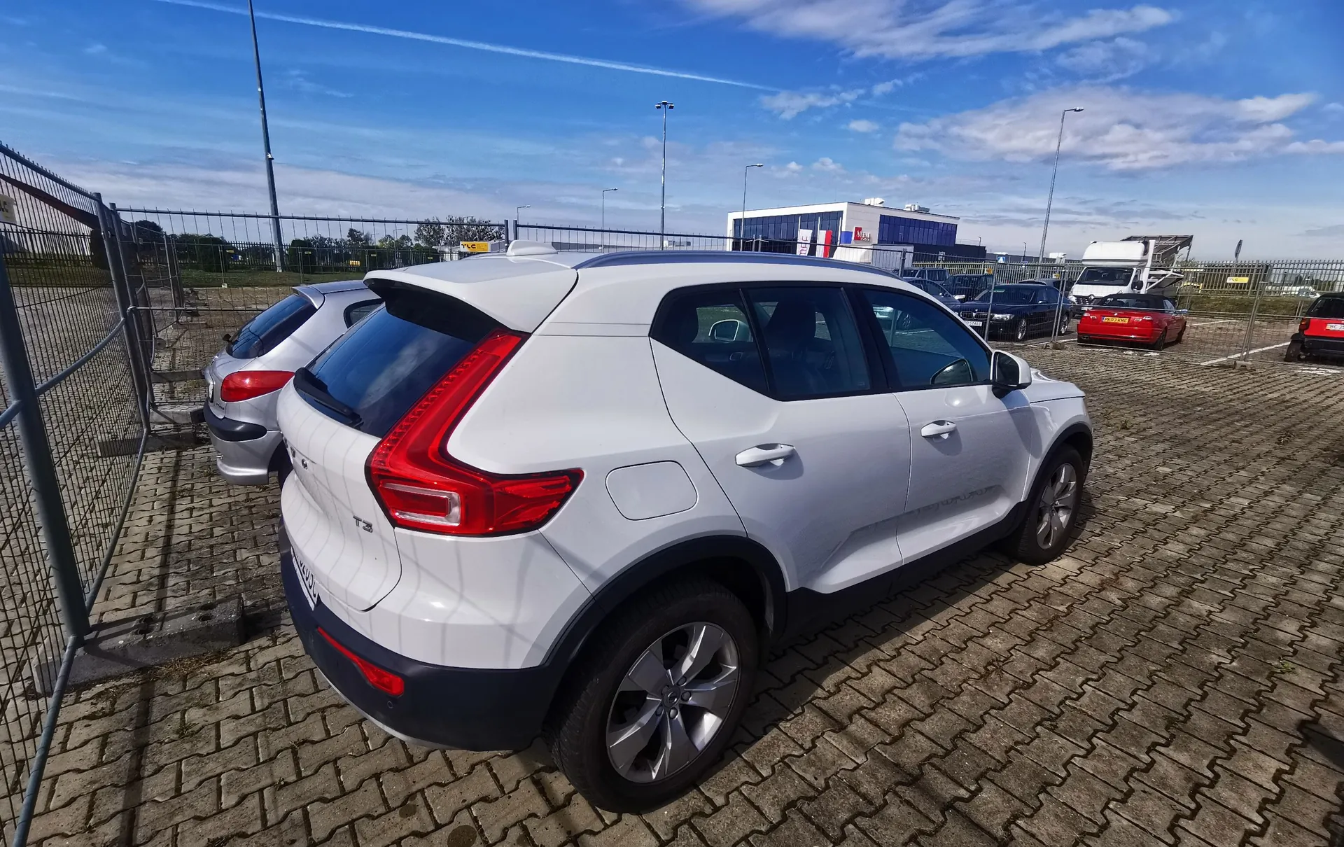 Biały samochód marki Volvo XC40 zaparkowany tyłem do obiektywu na placu wyłożonym kostką brukową. W tle widoczne są inne zaparkowane samochody, w tym srebrny hatchback po lewej stronie oraz czerwony samochód w oddali. W tle znajduje się niebieski budynek przemysłowy oraz błękitne niebo z delikatnymi smugami chmur. Po lewej stronie widoczny jest metalowy płot z siatki.