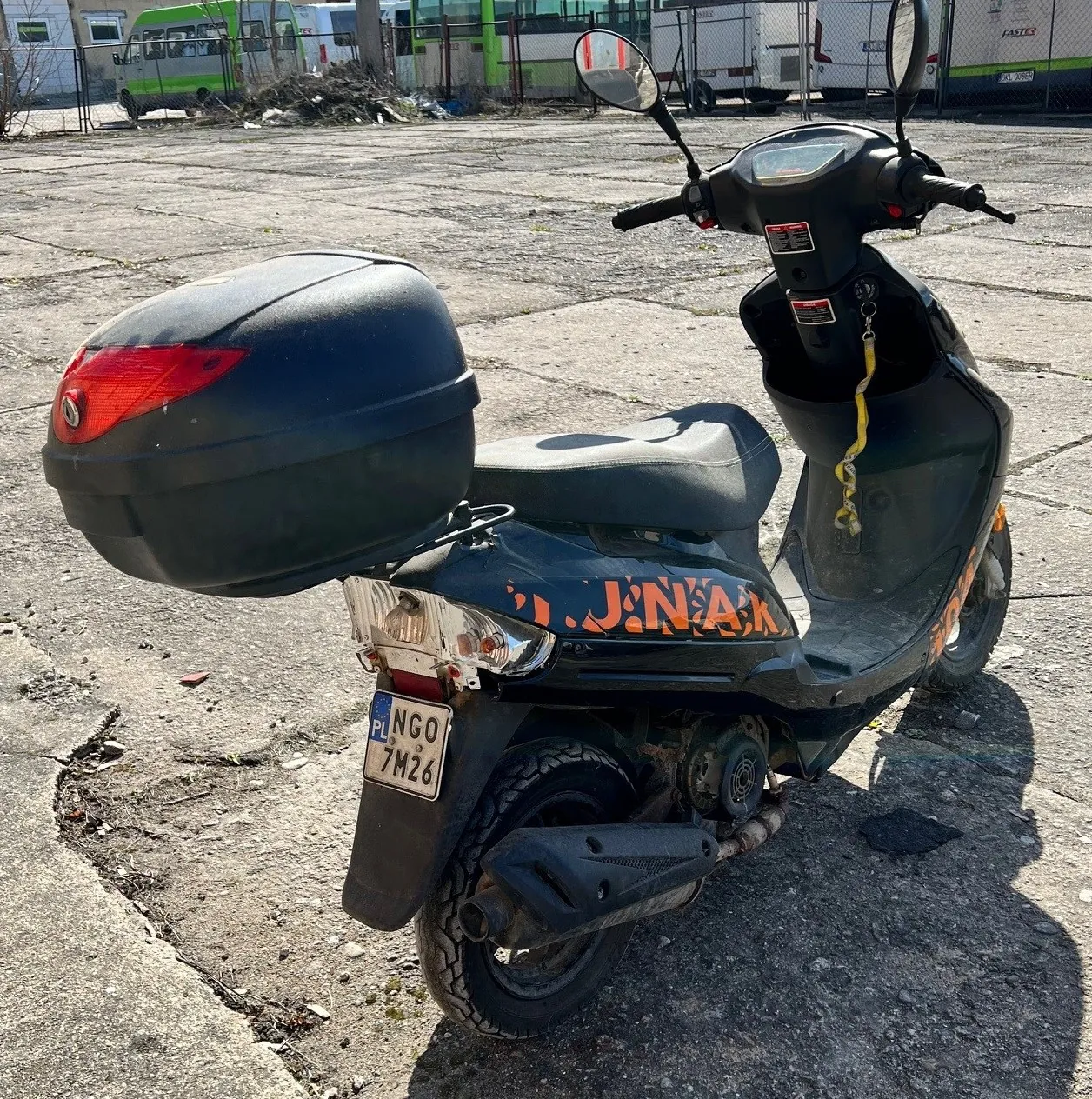 Czarny skuter z dużym czarnym bagażnikiem na tylnej części, czerwonym światłem tylnym i pomarańczowym napisem "J.N.A.K." na boku. Tablica rejestracyjna PL NGO 7M26. Skuter stoi na pękniętym betonie, w tle widoczne są zielono-białe autobusy i ogrodzenie. Na kierownicy znajdują się lusterka, a na przedniej części wiszący żółty pasek.