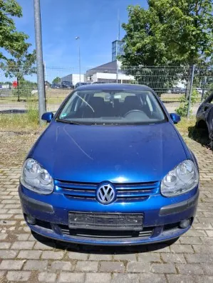 Niebieski samochód marki Volkswagen zaparkowany przodem do obiektywu na kostce brukowej. W tle widoczny jest metalowy płot, zielone drzewa oraz nowoczesny biały budynek pod jasnoniebieskim niebem.