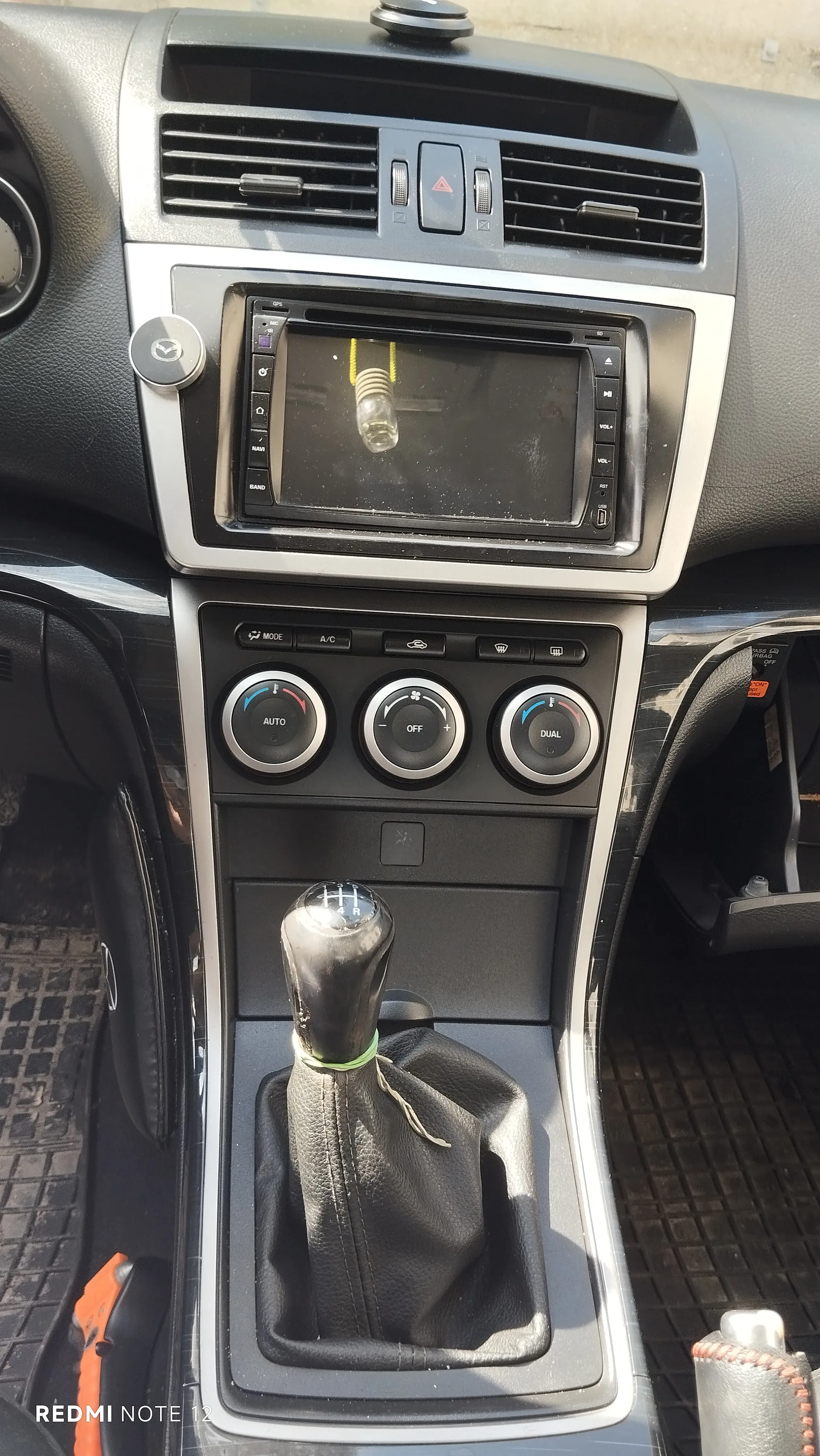 Wnętrze samochodu marki Mazda z widoczną centralną konsolą. Na konsoli znajduje się ekran multimedialny z odbiciem butelki z żółtym korkiem, trzy okrągłe przyciski klimatyzacji z napisami AUTO, OFF i DUAL, a poniżej dźwignia zmiany biegów z czarną obudową i zielonym paskiem. W lewym dolnym rogu widoczny jest napis REDMI NOTE 12. W tle widoczne są czarne deski rozdzielcze, klimatyzacyjne wywiewacze oraz przycisk awaryjny z czerwonym trójkątem.