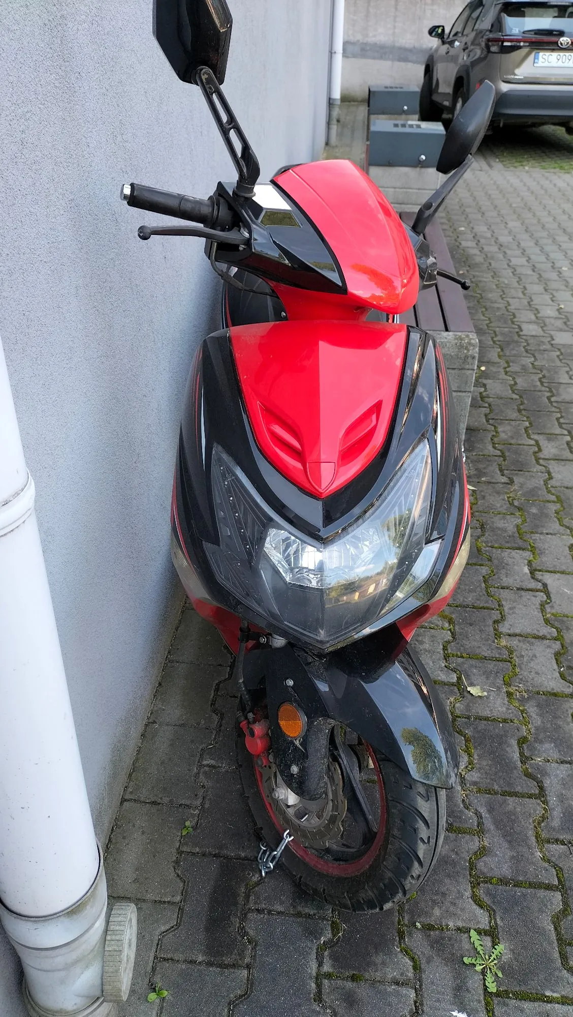 Czerwono-czarny skuter zaparkowany na kostce brukowej przy szarej ścianie budynku. Pojazd jest ustawiony przodem do obiektywu. W tle widoczny jest fragment srebrnego samochodu z tablicą rejestracyjną zaczynającą się od liter SC.