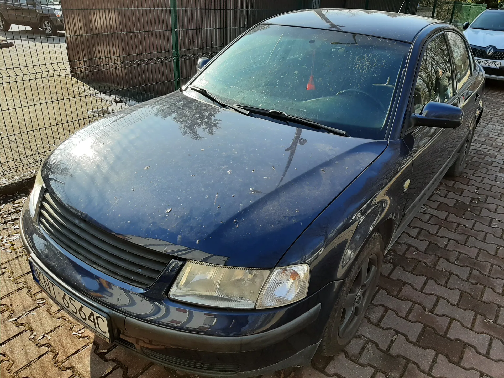 Ciemnoniebieski samochód osobowy marki Audi zaparkowany na brukowanej powierzchni. Widoczna tablica rejestracyjna KNT 6564C na przodzie. W tle biały samochód z tablicą KNT 8875A, metalowe ogrodzenie, brązowa budowla i resztki śniegu na ziemi. Na masce samochodu znajdują się drobne zanieczyszczenia. Oświetlenie słoneczne, cień pada na lewą stronę pojazdu.