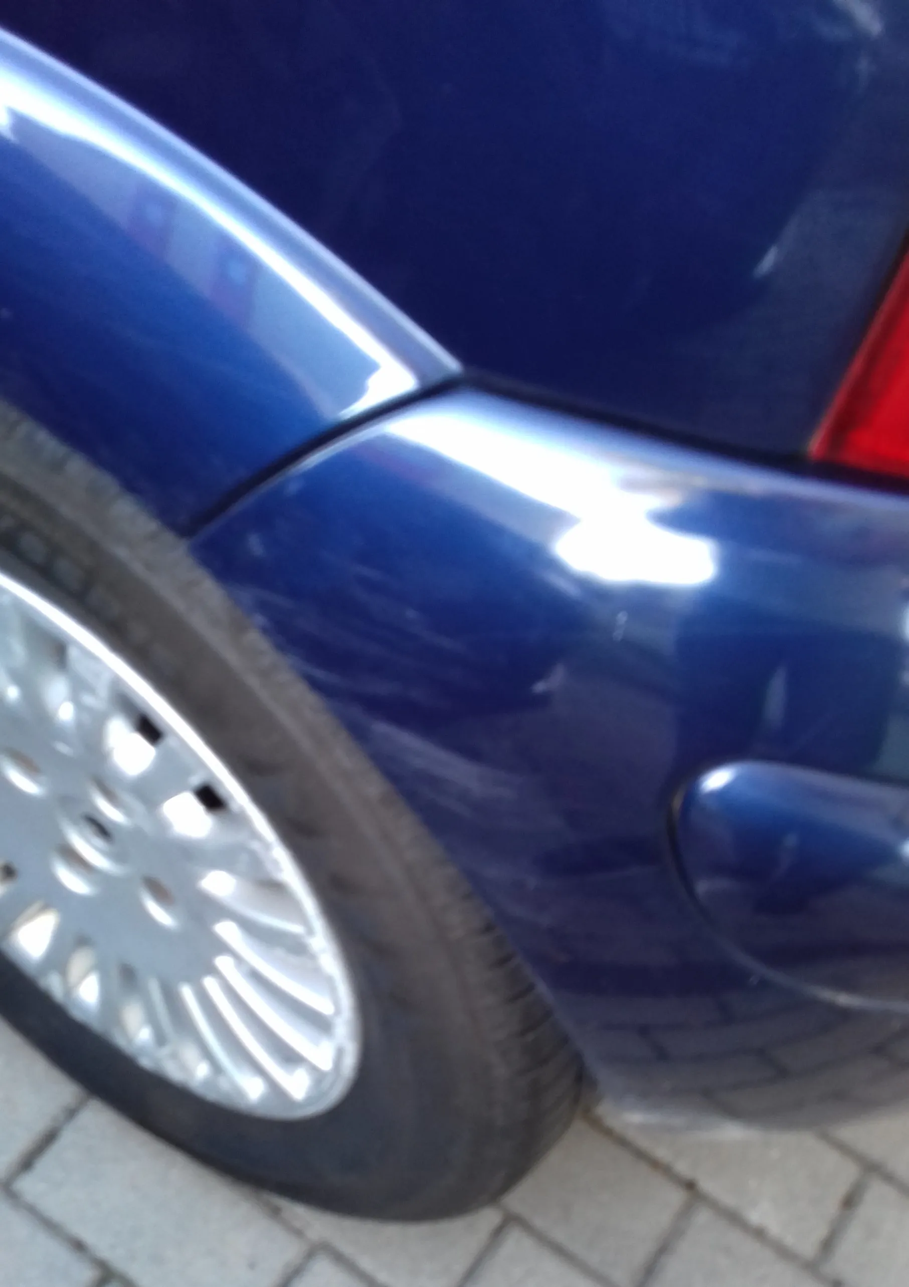 Zbliżenie na tylny prawy błotnik i koło ciemnoniebieskiego samochodu. Widoczna jest część opony z felgą aluminiową oraz fragment tylnej lampy w prawym górnym rogu.