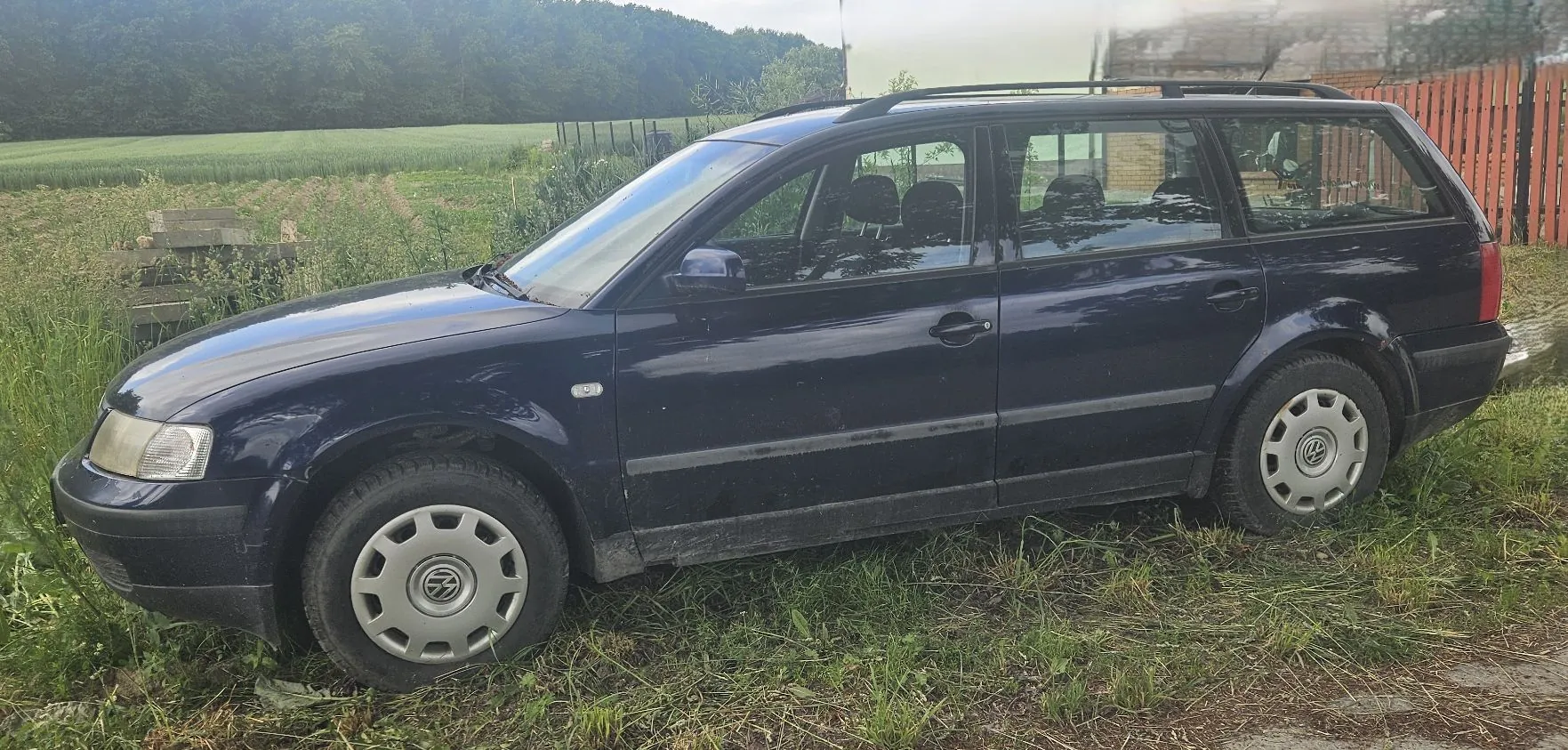 Ciemnoniebieski samochód marki Volkswagen w nadwoziu kombi, zaparkowany na trawiastym poboczu przy polu. Pojazd jest widoczny z profilu, posiada srebrne felgi z logo producenta oraz relingi dachowe. W tle widać zielone pole, las oraz fragment drewnianego ogrodzenia.