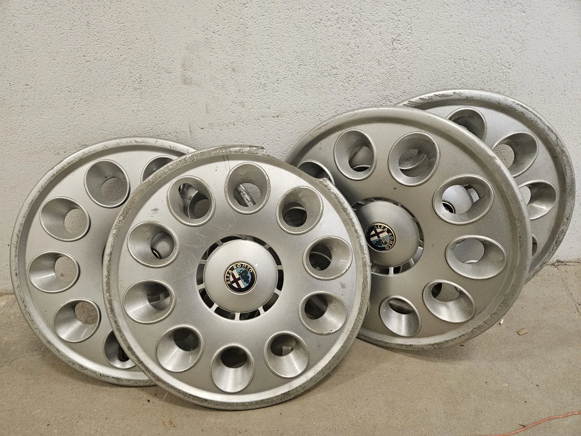 Cztery srebrne, aluminiowe felgi samochodowe o charakterystycznym wzorze z wieloma okrągłymi otworami, oparte o jasną, chropowatą ścianę. Na środku każdej felgi znajduje się okrągły emblemat z logo marki Toyota.