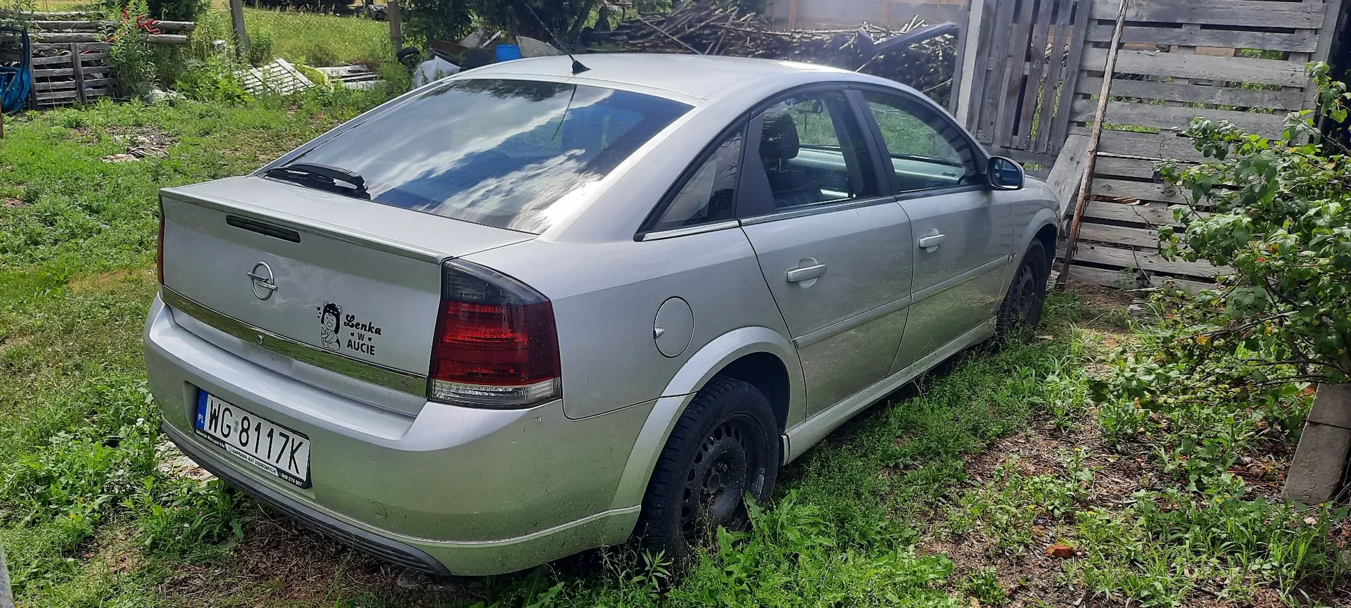 Srebrny samochód osobowy marki Opel zaparkowany na trawiastym terenie obok drewnianego ogrodzenia. Na klapie bagażnika znajduje się naklejka z napisem „Lenka w aucie” oraz polska tablica rejestracyjna o numerze WG 8117K.