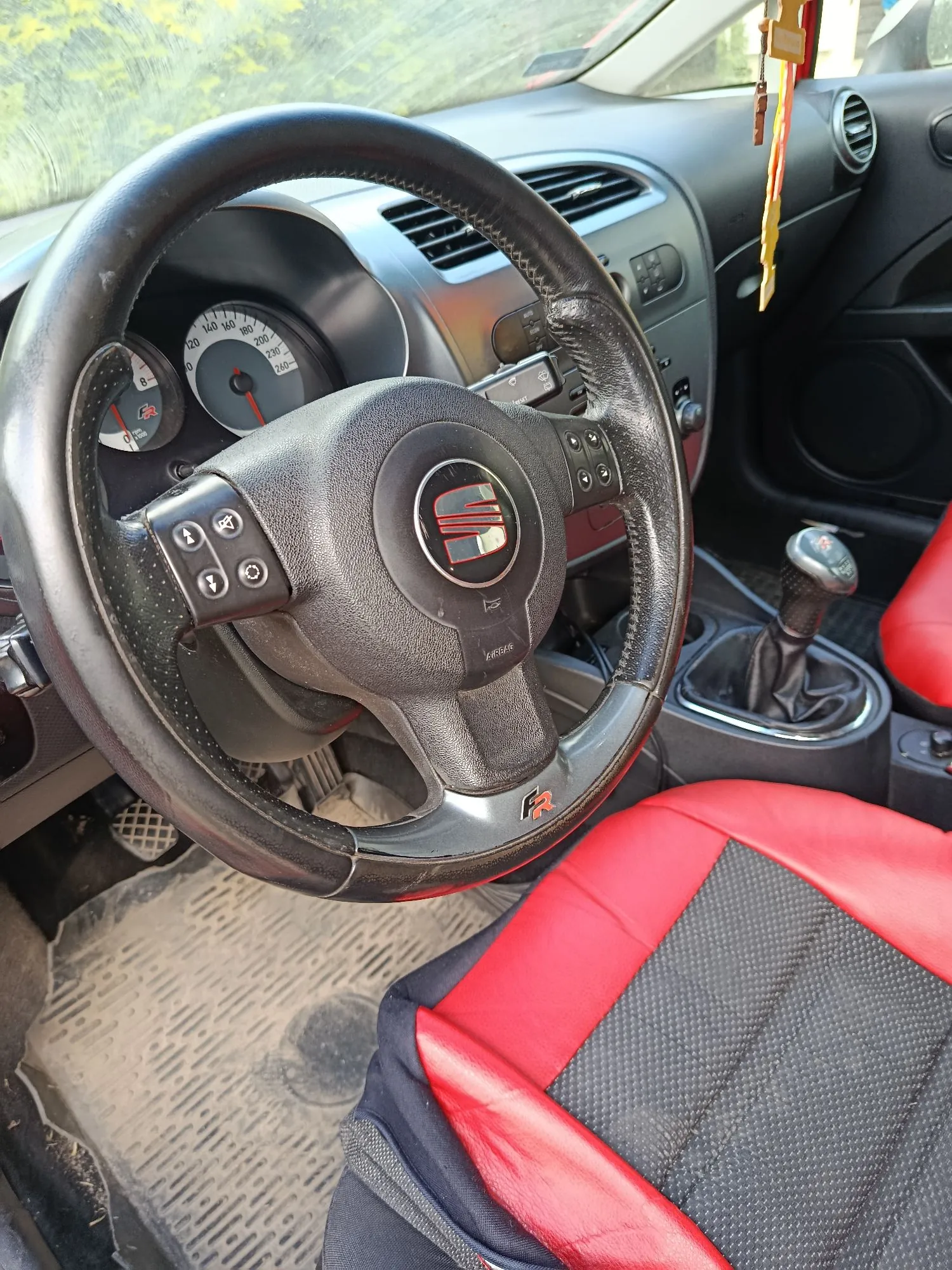 Wnętrze samochodu marki SEAT z perspektywy kierowcy. Widoczna jest czarna, skórzana kierownica z logo SEAT na środku i oznaczeniem 'FR' na dolnym ramieniu. W tle znajduje się deska rozdzielcza z zegarami, kratkami nawiewów oraz lewarem zmiany biegów. Fotel kierowcy jest dwukolorowy: czerwony i czarny. Na podłodze widoczna jest gumowa mata.